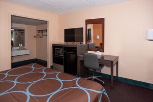 Una habitación de hotel con cama, escritorio, televisor, espejo y kitchenette.