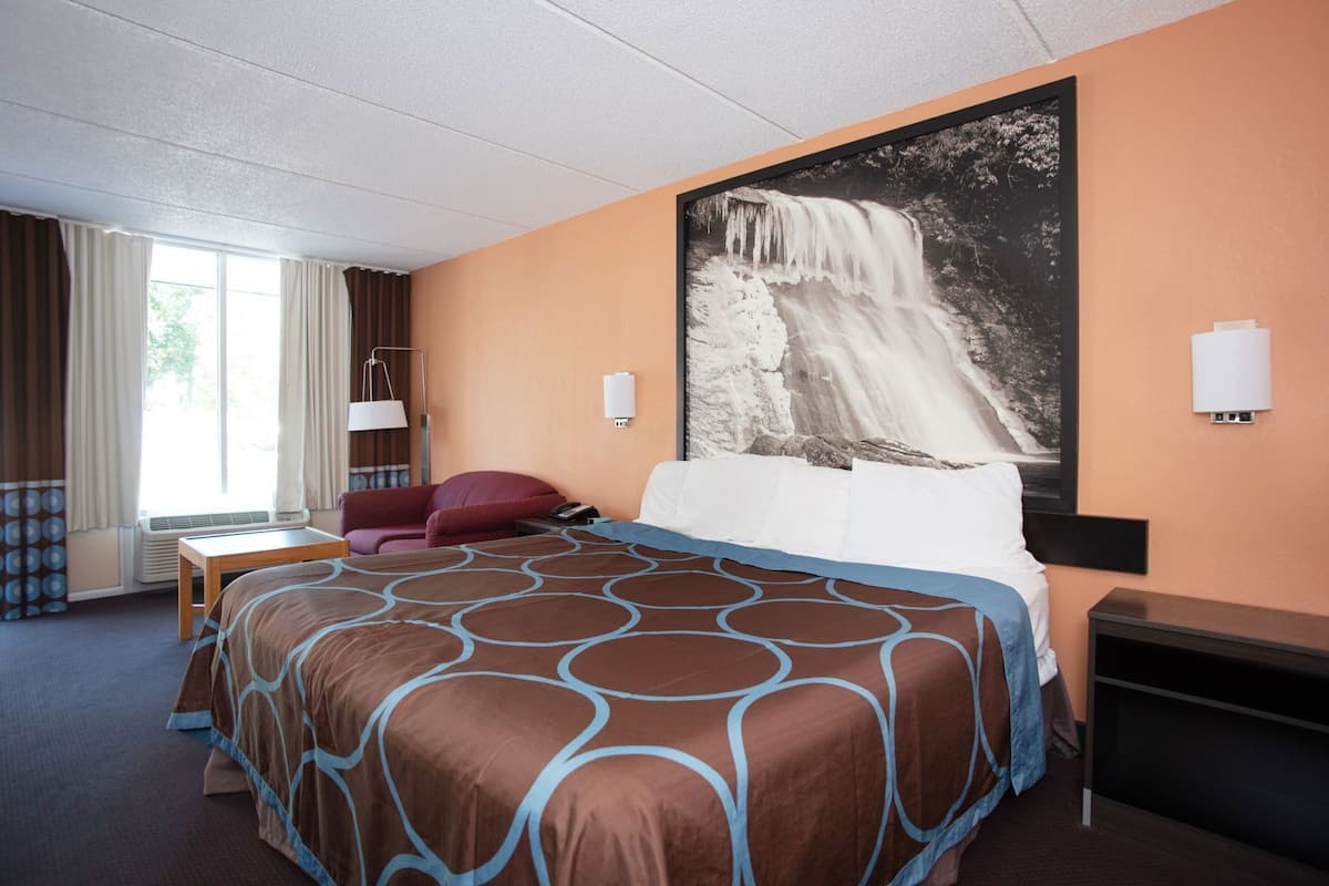 Ett hotellrum med en stor säng, ett skrivbord, en stol, ett fönster med gardiner och en vägglampa.