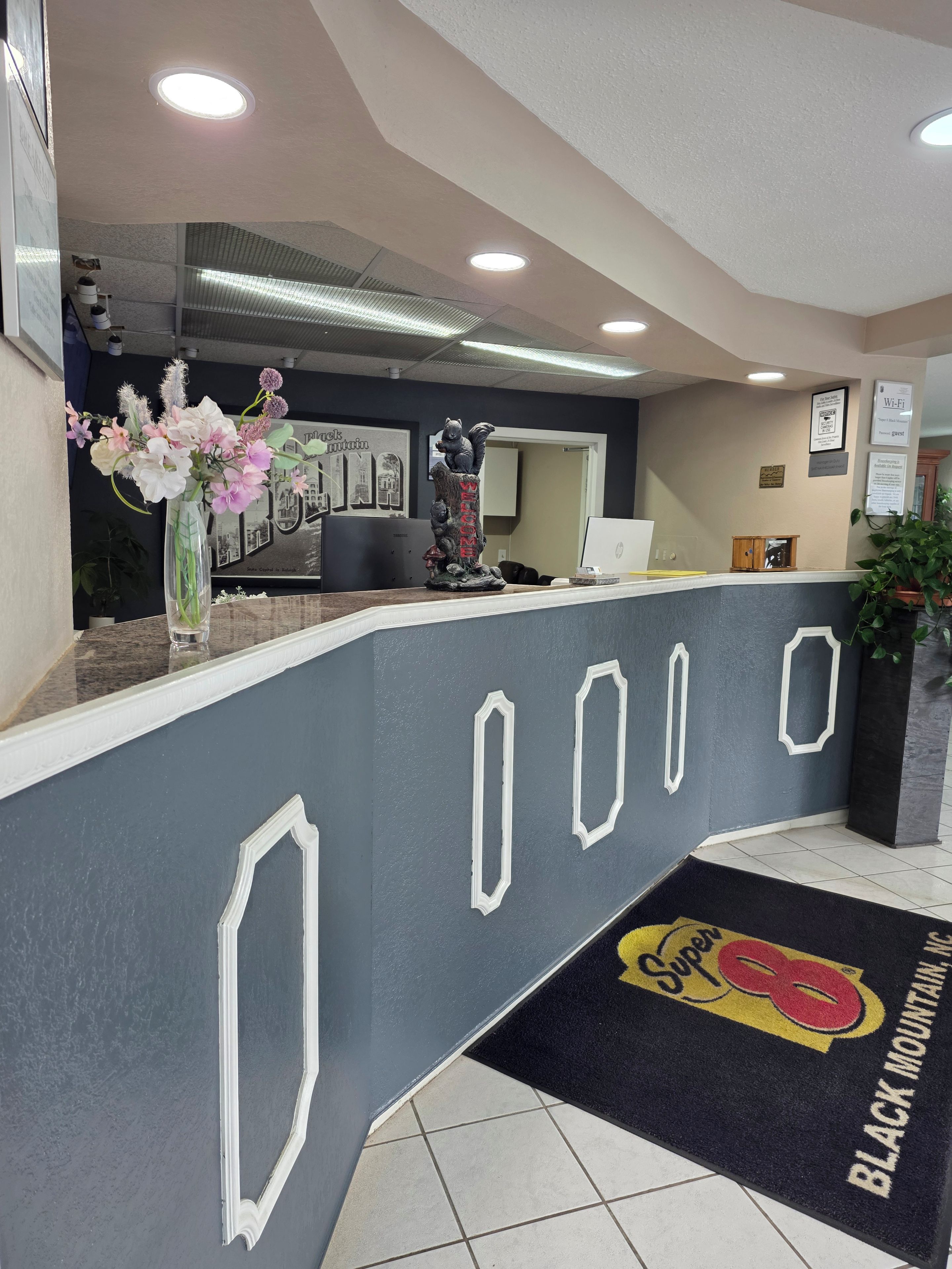A reception desk with a vase of flowers, a framed picture, and a doormat with the hotel's name.