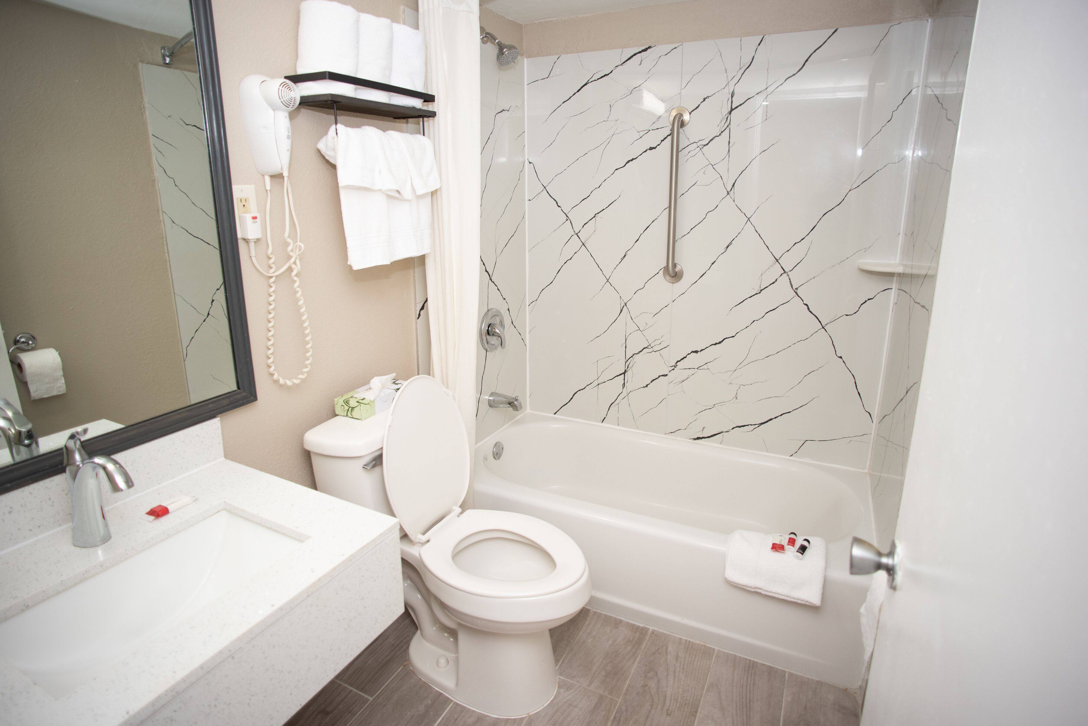 Un baño con lavamanos, inodoro y bañera blancos. Hay un secador de pelo en la pared y un perchero con toallas.