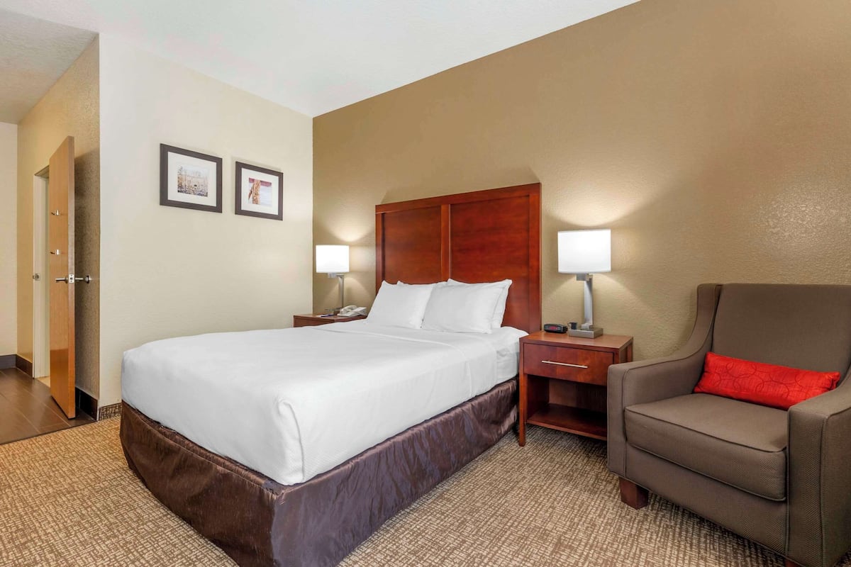 Quarto de hotel com uma cama grande, duas luminárias de cabeceira, uma mesa de cabeceira de madeira, uma poltrona marrom com almofada vermelha e duas fotos emolduradas na parede.