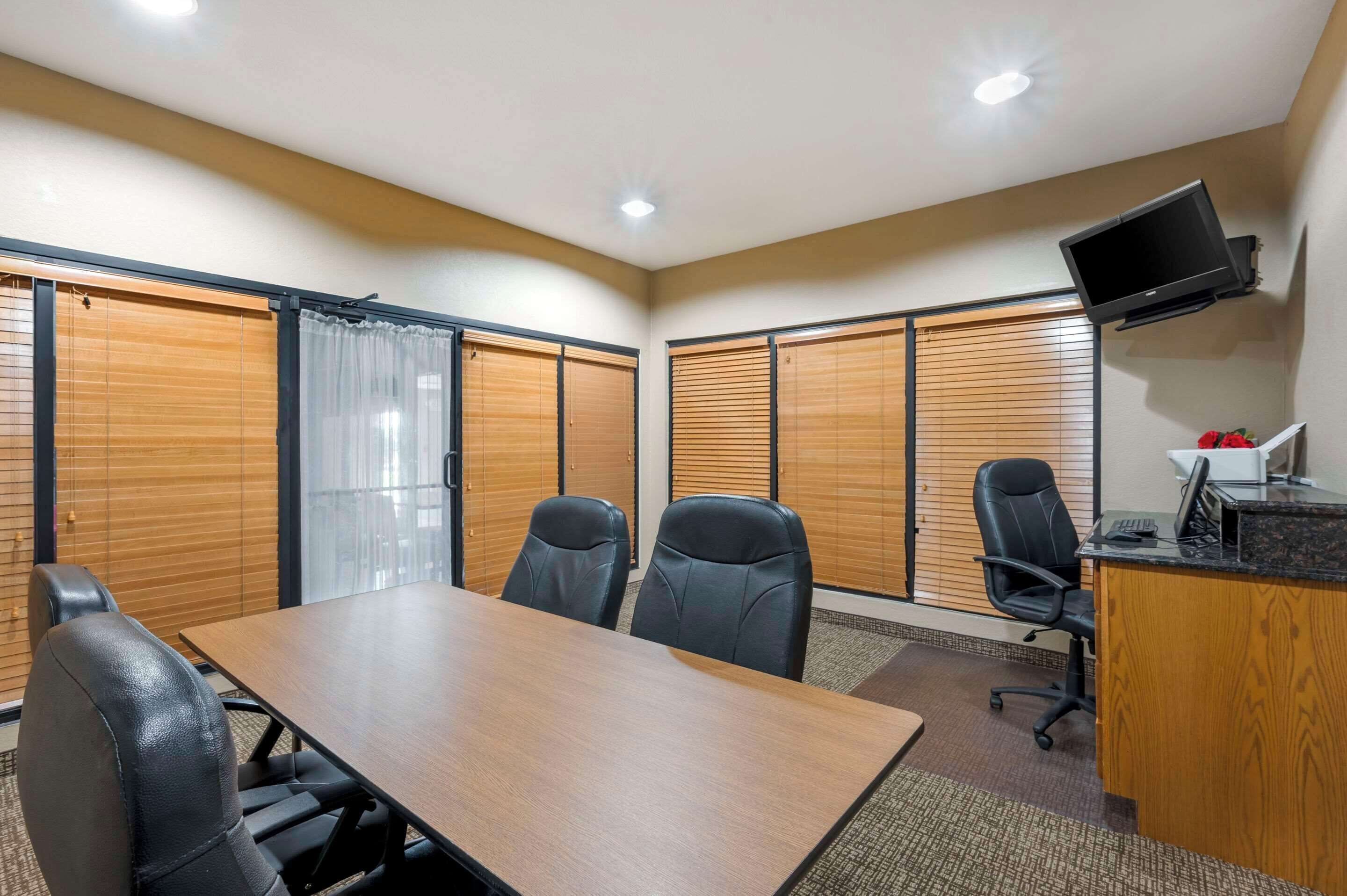 Uma sala de conferências com uma mesa comprida, quatro cadeiras, uma escrivaninha com computador e uma televisão fixada na parede.