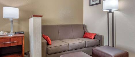 A hotel room with a sofa, two ottomans, a desk with a lamp, and a floor lamp.