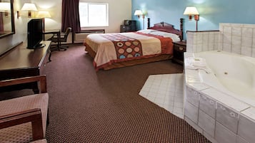 A hotel room with a bed, a desk, a chair, a TV, a desk lamp, a bedside table with a lamp, and a bathtub.