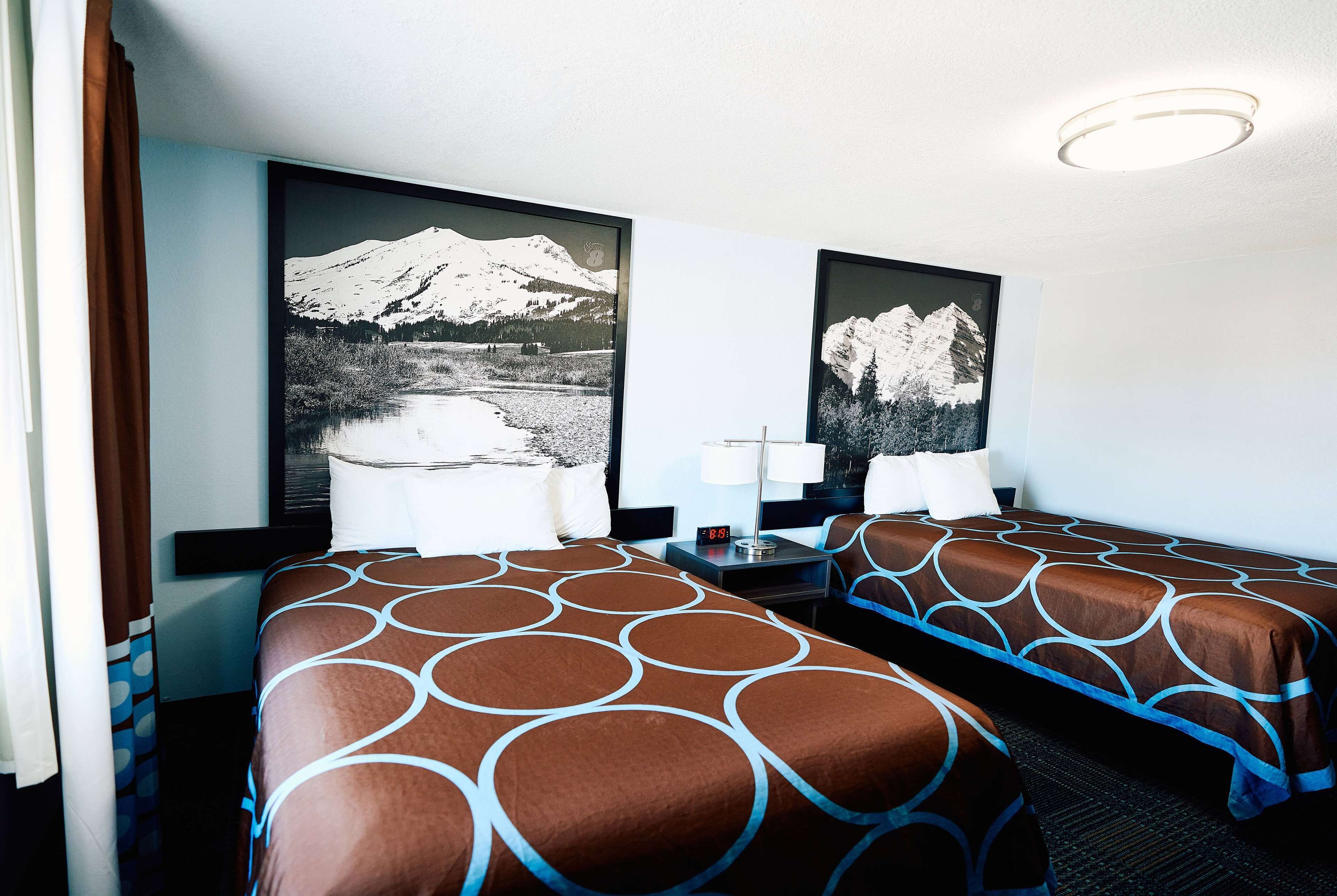 Habitación de hotel con dos camas, una mesita de noche, una lámpara y dos cuadros con paisajes de montañas.