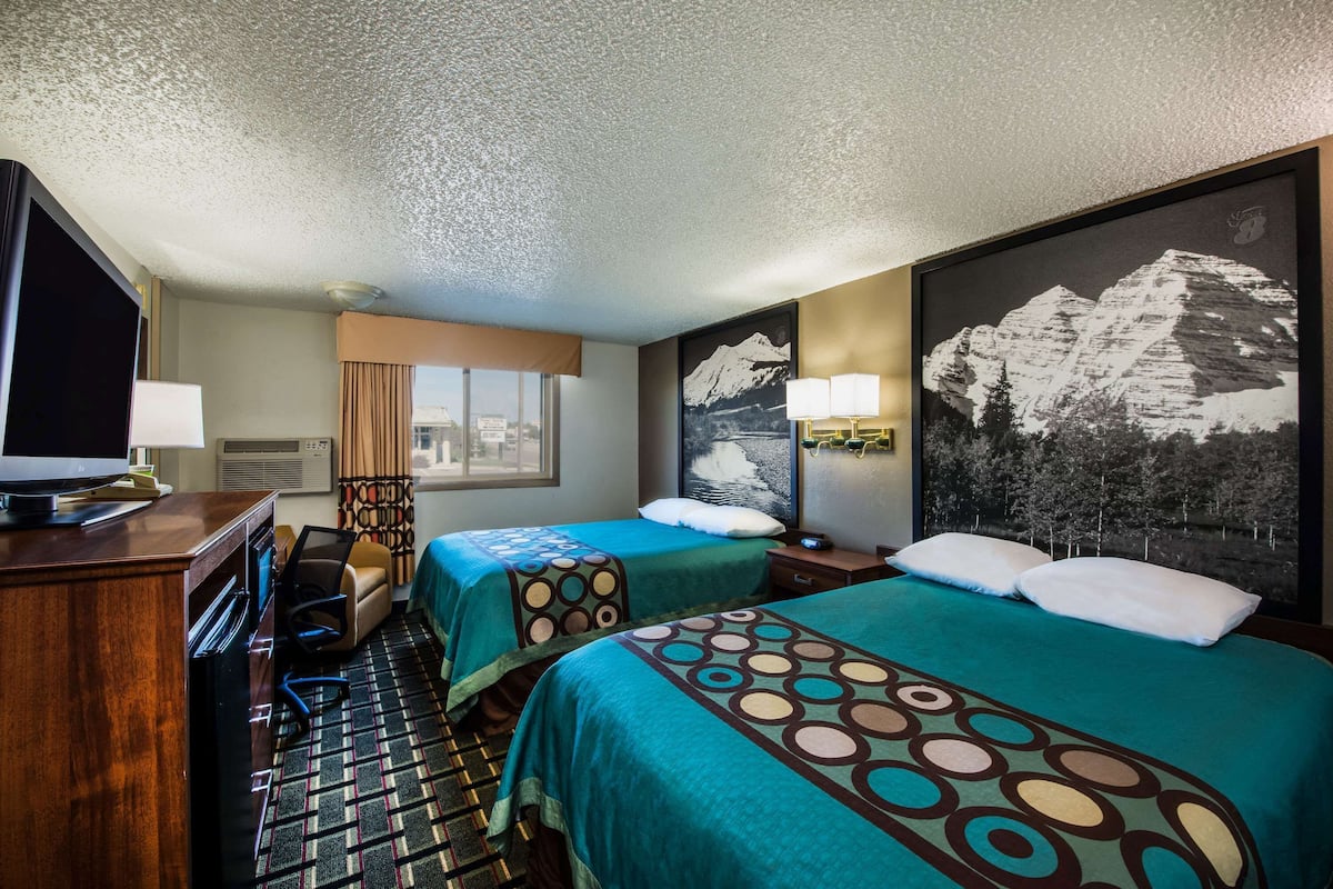 Habitación de hotel con dos camas, un escritorio con televisor, una ventana con cortinas y un cuadro con vista a la montaña.