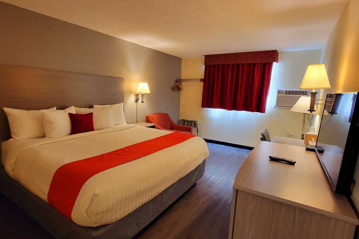 Hotelkamer met een groot bed, twee nachtkastjes, een bureau met een stoel, een spiegel en een raam met rode gordijnen.