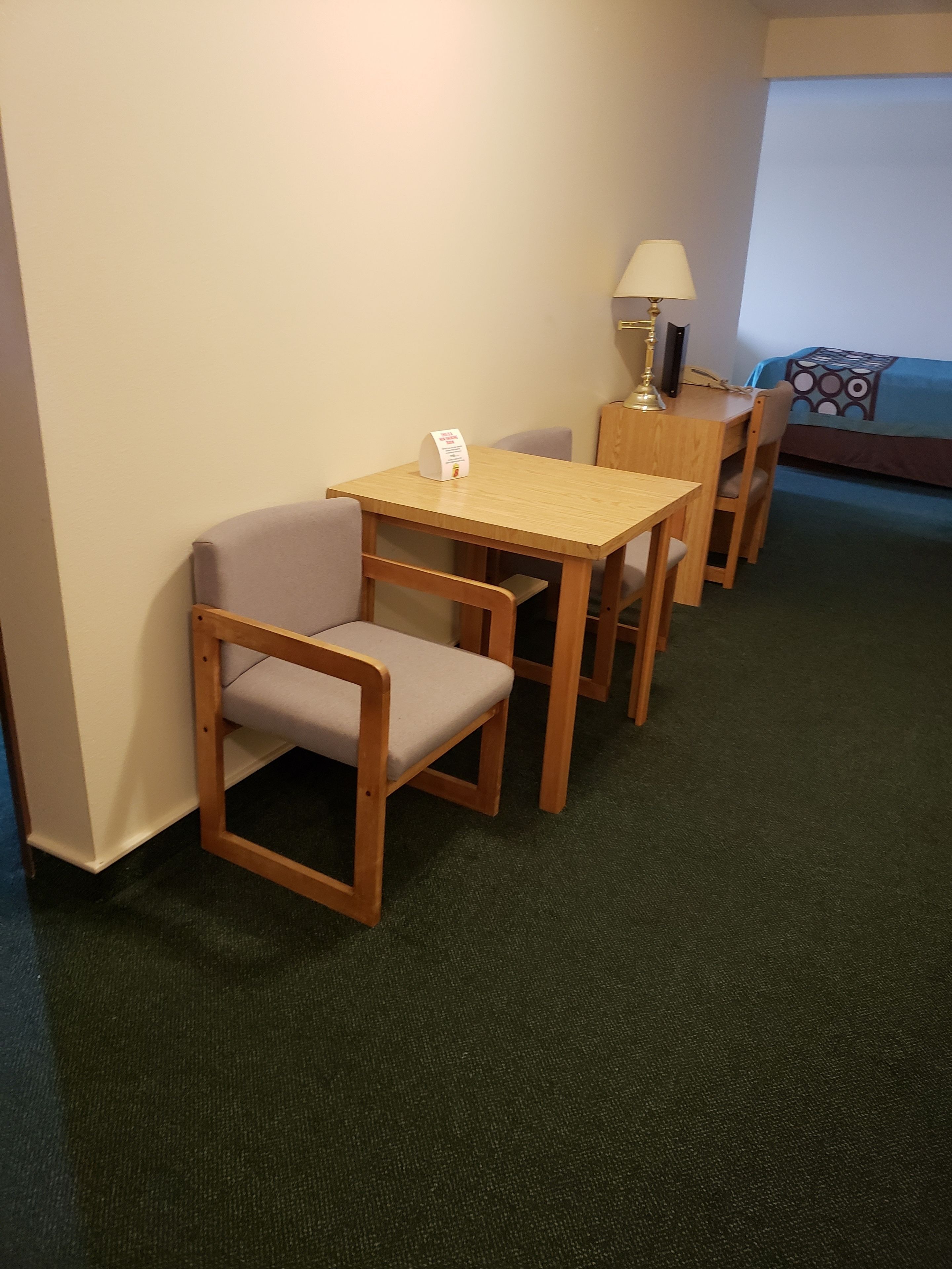 Une chambre d’hôtel avec une table et des chaises en bois, une lampe, un lit recouvert d’une couvre-lit à motifs et un tapis vert.