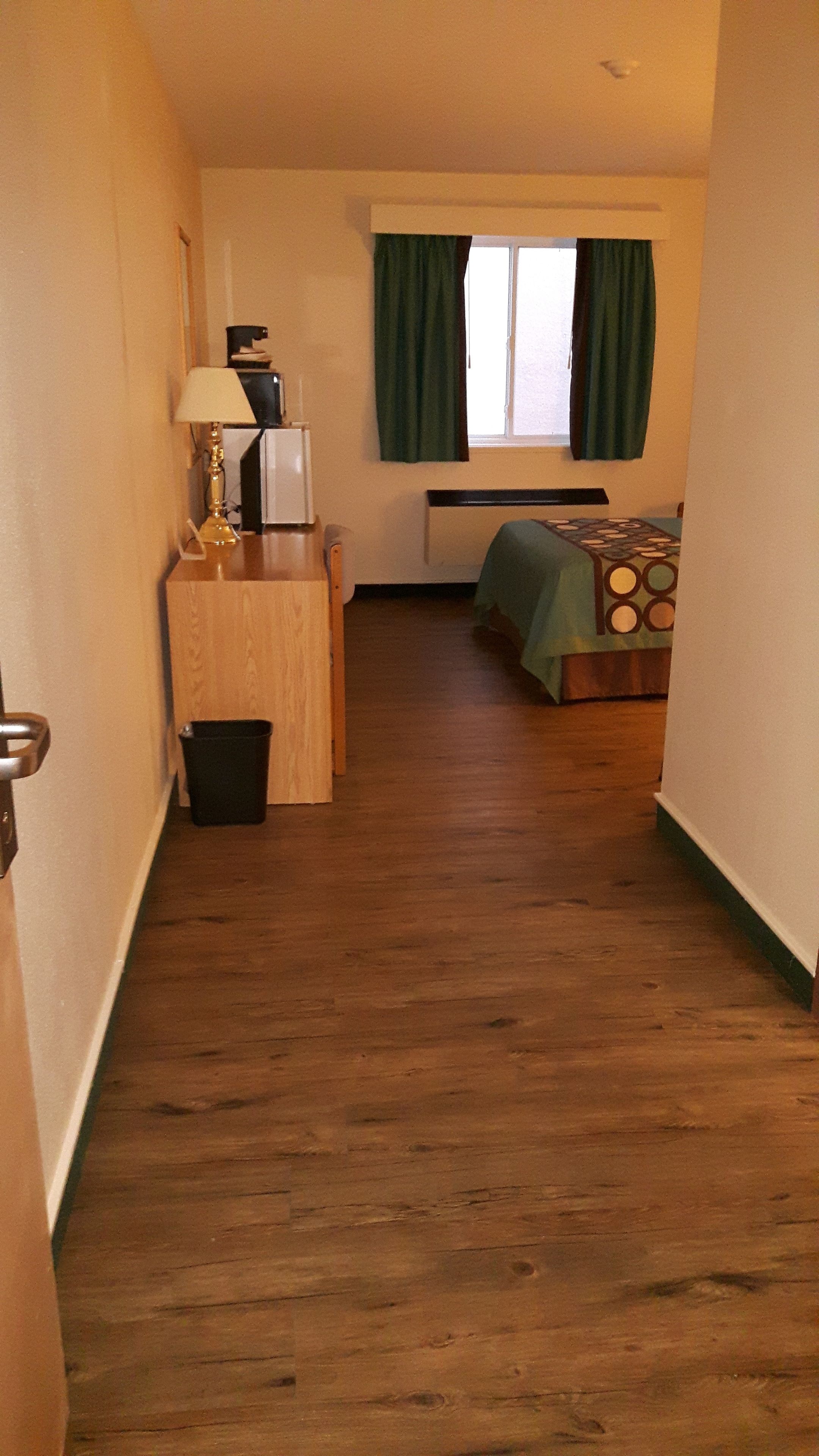 Un estrecho pasillo con suelo de madera que conduce a una habitación con una cama, un escritorio, una lámpara y una ventana con cortinas.