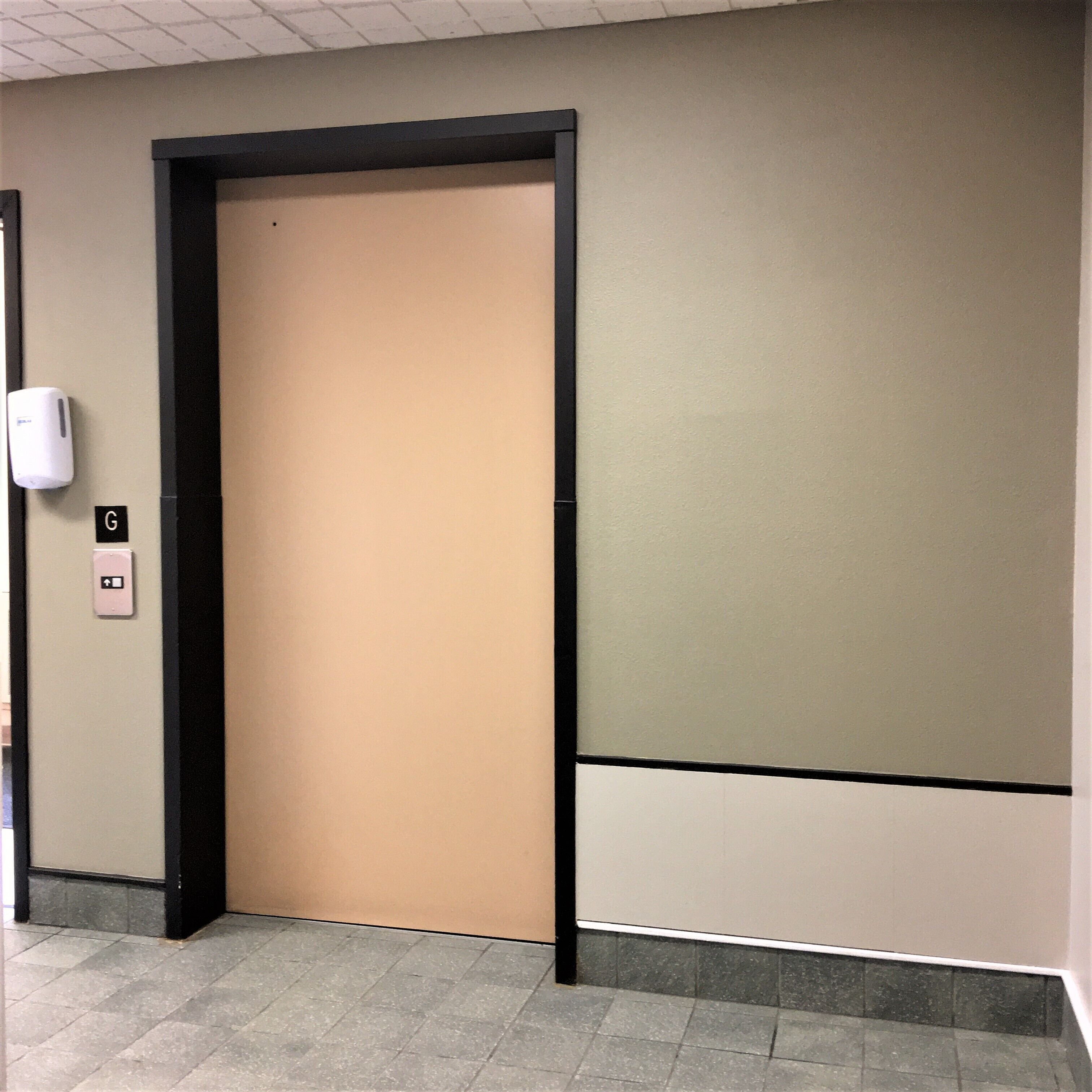 A hallway with a beige elevator door, a hand sanitizer dispenser, and a sign with an arrow pointing left.
