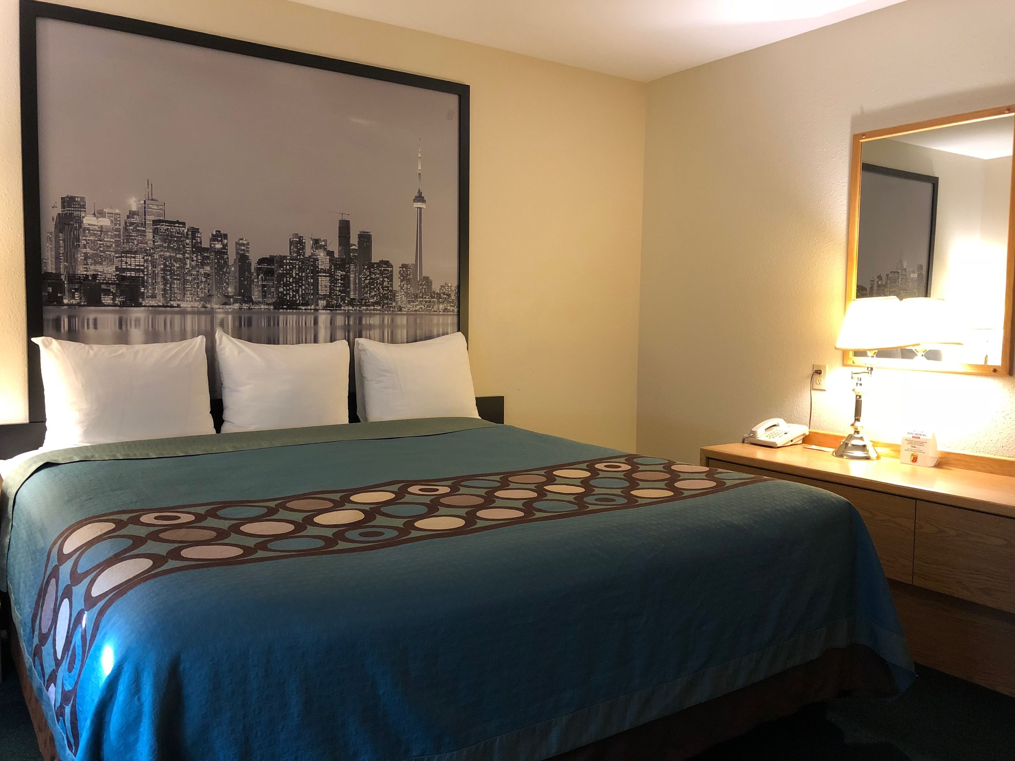 Ein Hotelzimmer mit einem Bett, einer Stadtansicht als Wandbild, einer Nachttischlampe und einem Nachttisch mit Telefon und Taschentuchbox.
