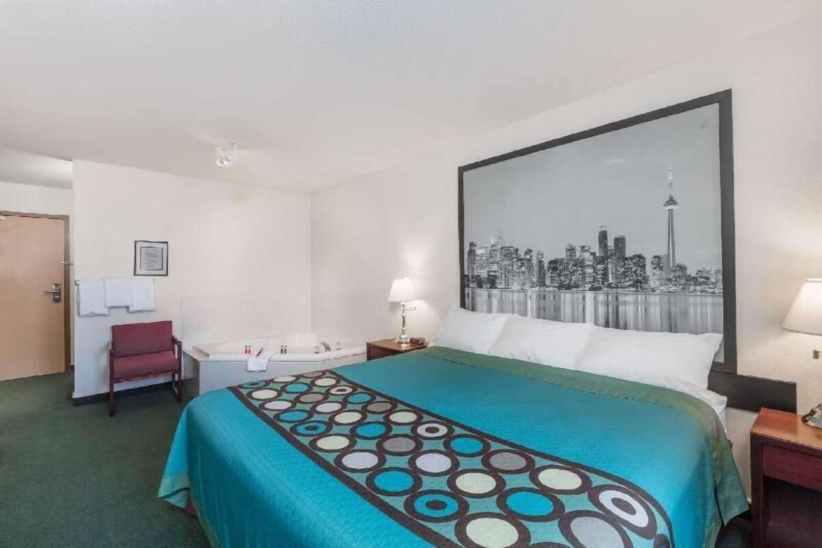 Habitación de hotel con cama, una silla, un escritorio, una lámpara y un cuadro grande con una vista urbana en la pared.