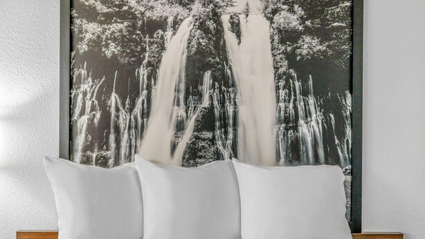 Una cama con ropa de cama y almohadas blancas, un cabecero de madera y un cuadro grande de una cascada en blanco y negro en la pared de fondo.