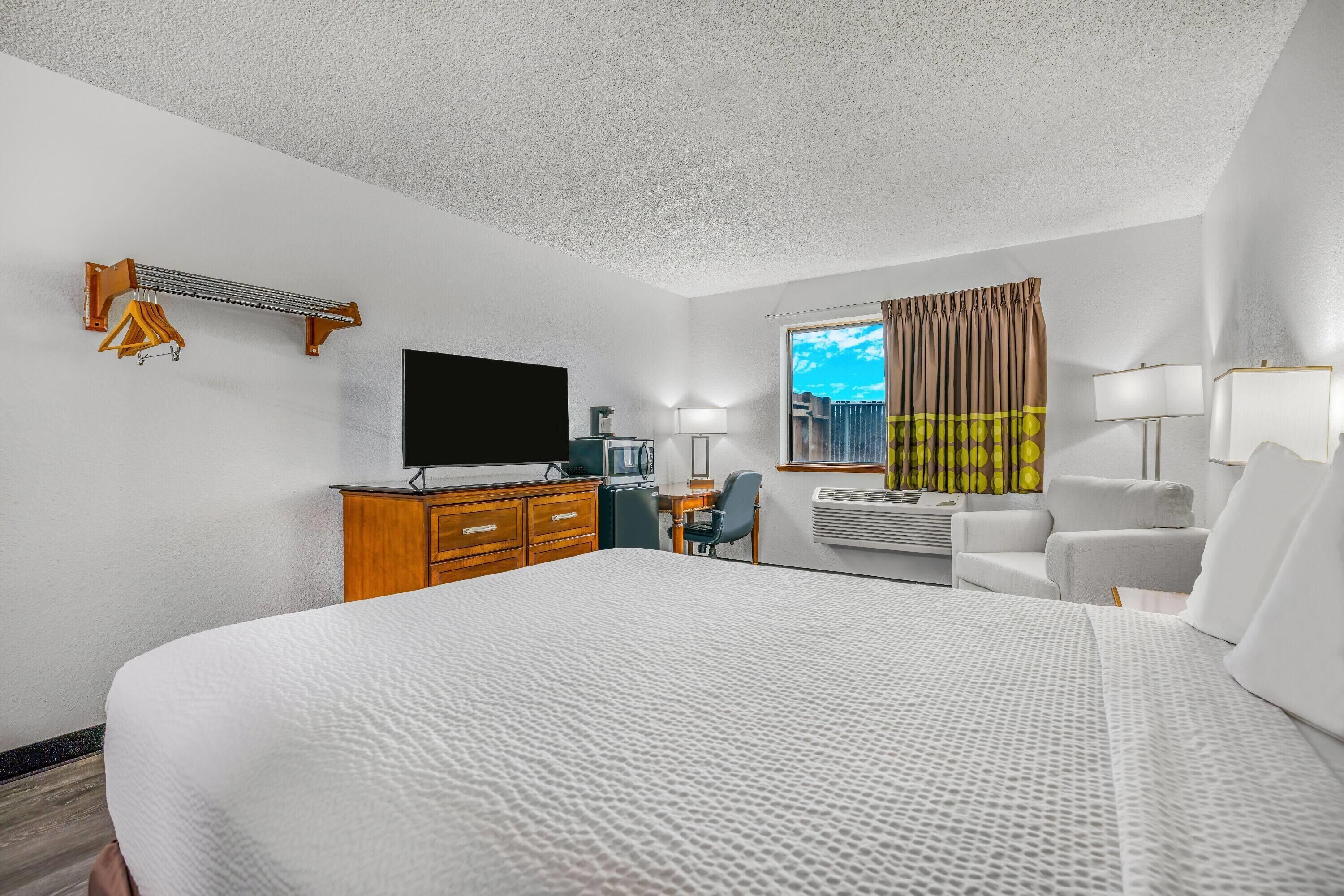 A hotel room with a large bed, a desk with a chair, a television, a window with curtains, and a wall-mounted heater.