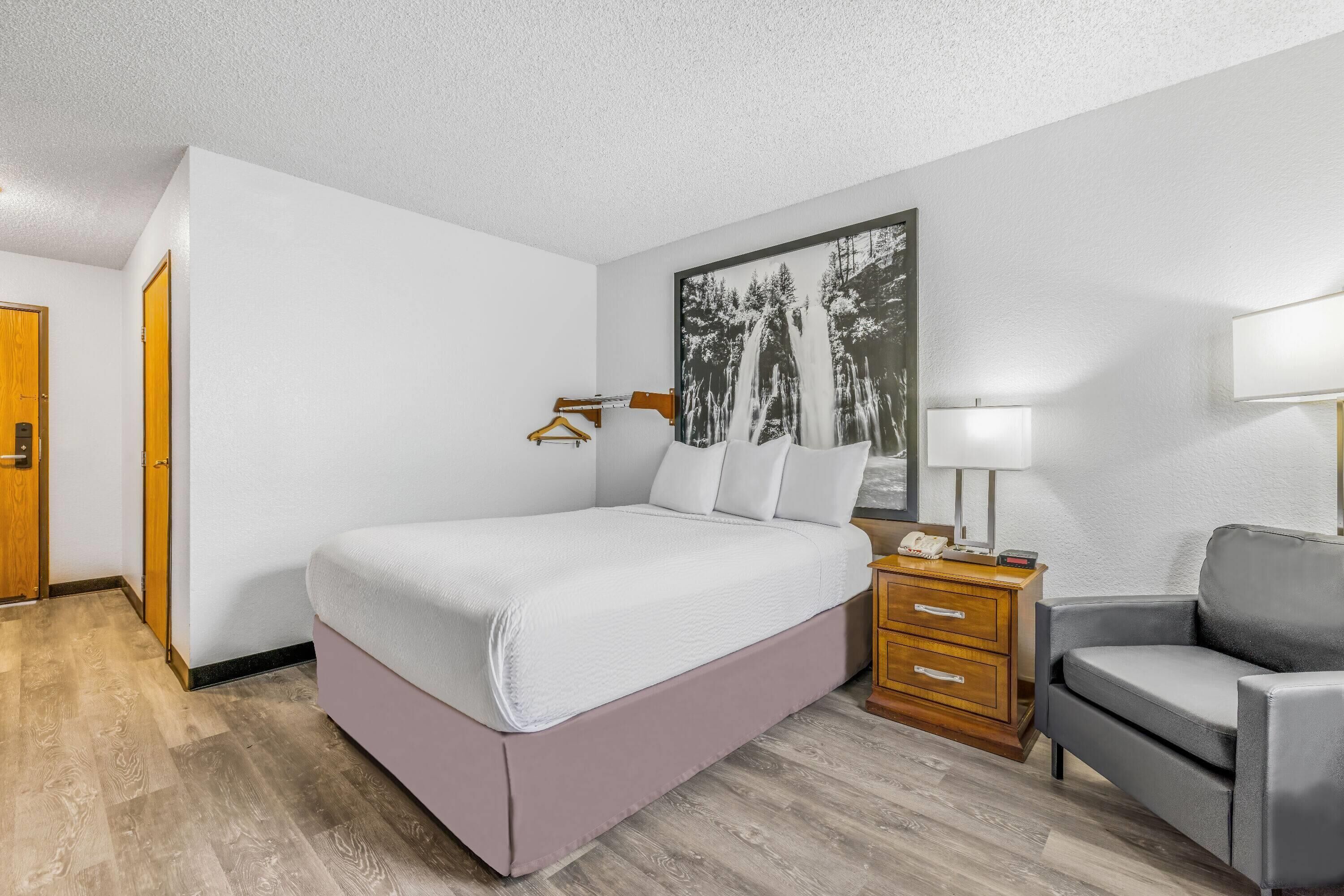 A hotel room with a large bed, a wooden nightstand, a gray armchair, and a wall-mounted lamp.