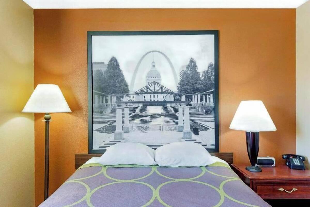 Une chambre d’hôtel avec un lit, deux lampes de chevet, une table de chevet avec un téléphone et un tableau encadré représentant un bâtiment accroché au mur.