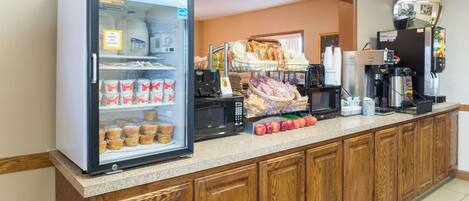 Un espace où l'on peut se servir soi-même pour le petit-déjeuner, comprenant un réfrigérateur à boissons, une machine à café et divers aliments disposés sur le comptoir.