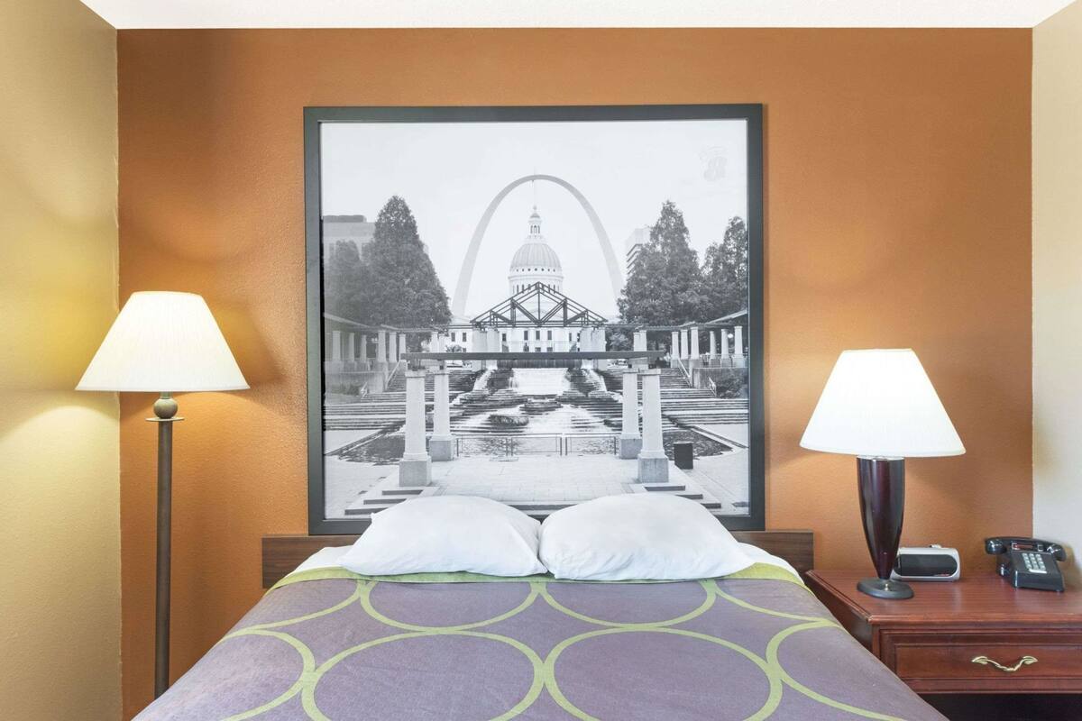 Une chambre d’hôtel avec un lit, deux lampes de chevet, un tableau encadré représentant un bâtiment, et une table de chevet avec un téléphone et une télécommande.