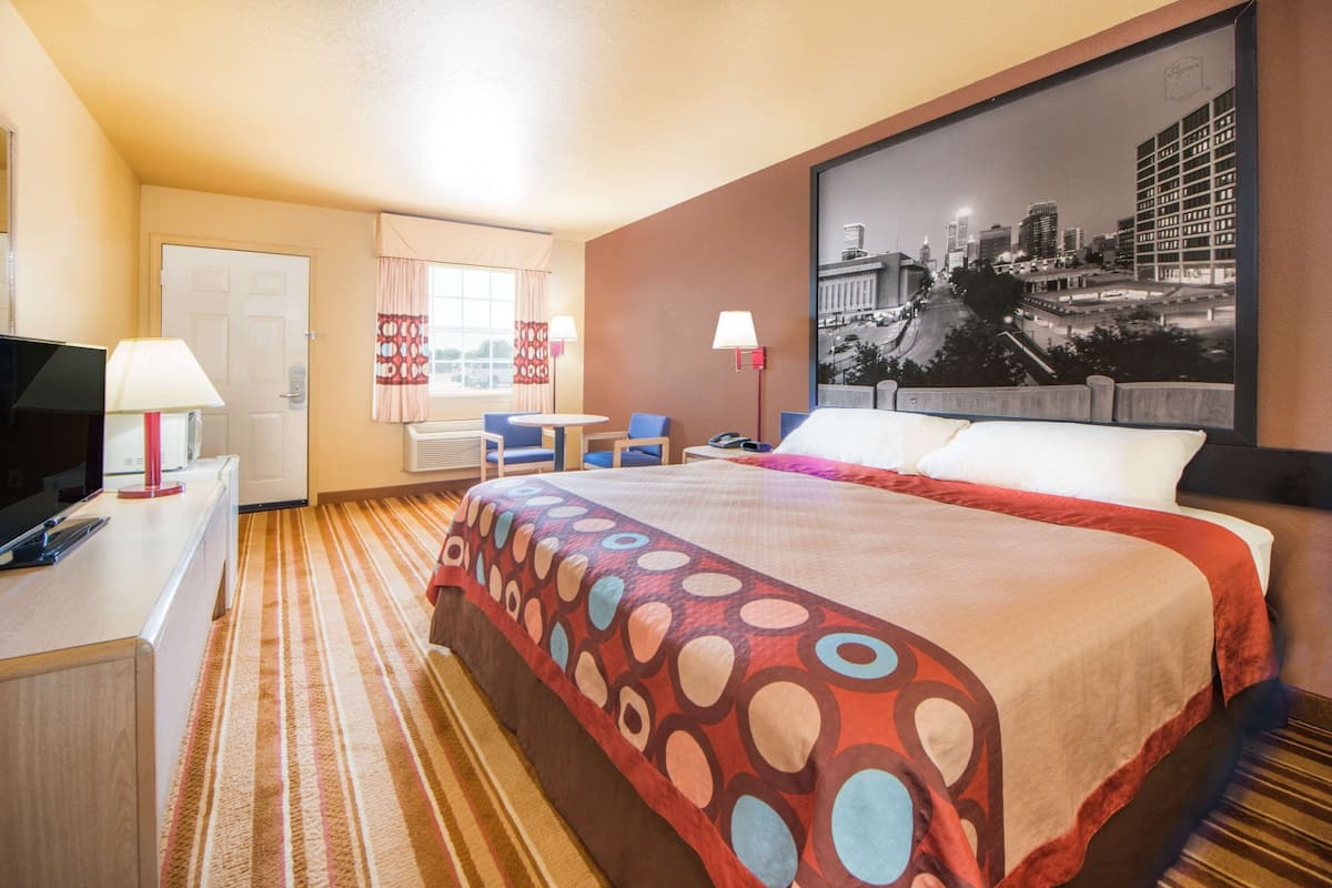 A hotel room with a large bed, a desk with a TV, a chair, a small table, and a window with curtains.