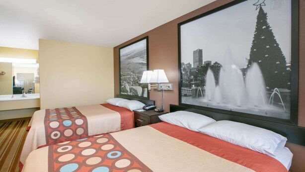 A hotel room with two beds, a large window with a cityscape view, a nightstand with a lamp, and a framed picture of a fountain.