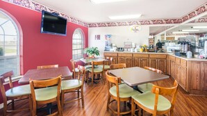 A restaurant with wooden tables and chairs, a TV mounted on the wall, and a kitchen area visible through an open doorway.