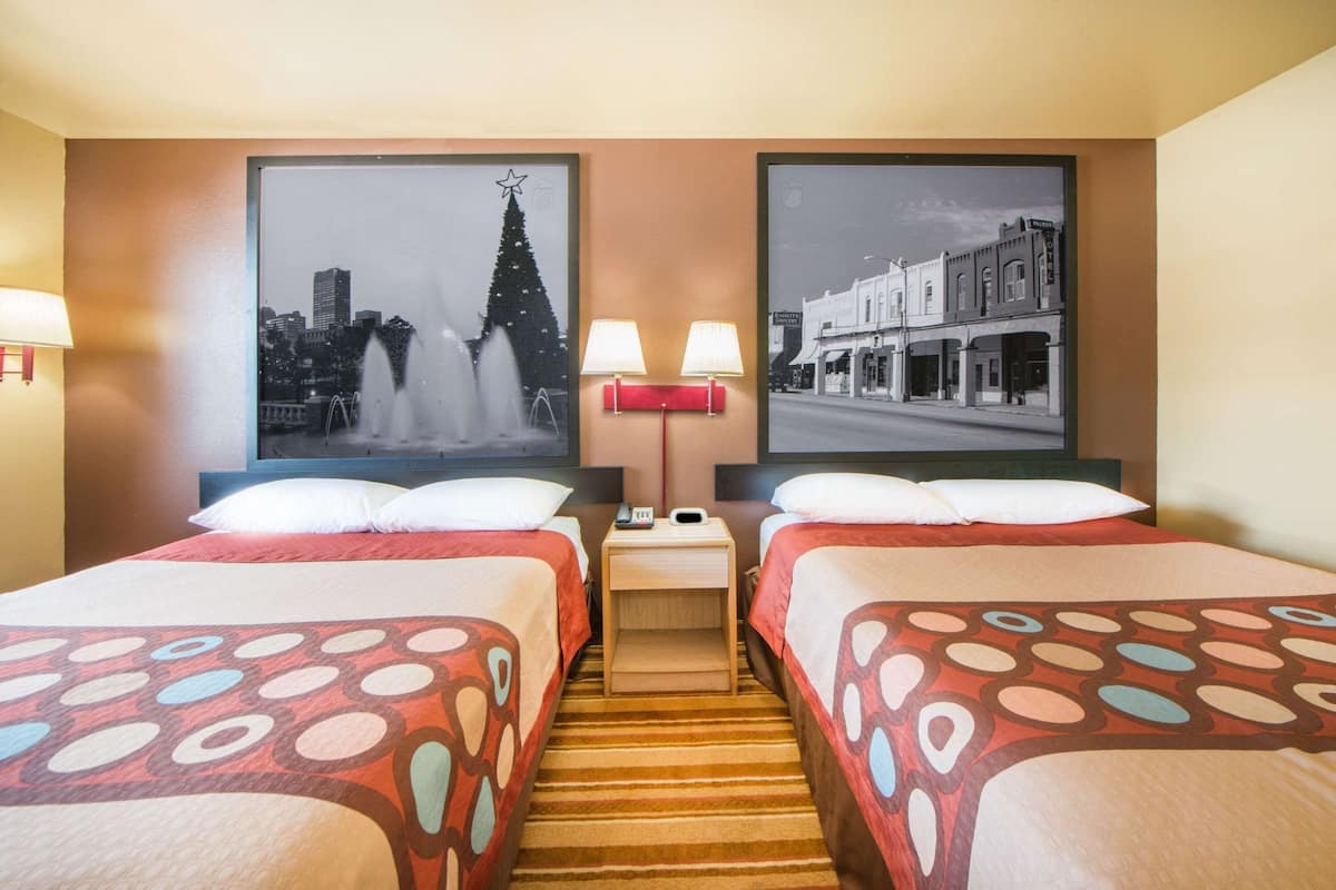 A hotel room with two beds, a nightstand with lamps, a cityscape picture on the wall, and a view of a street scene.