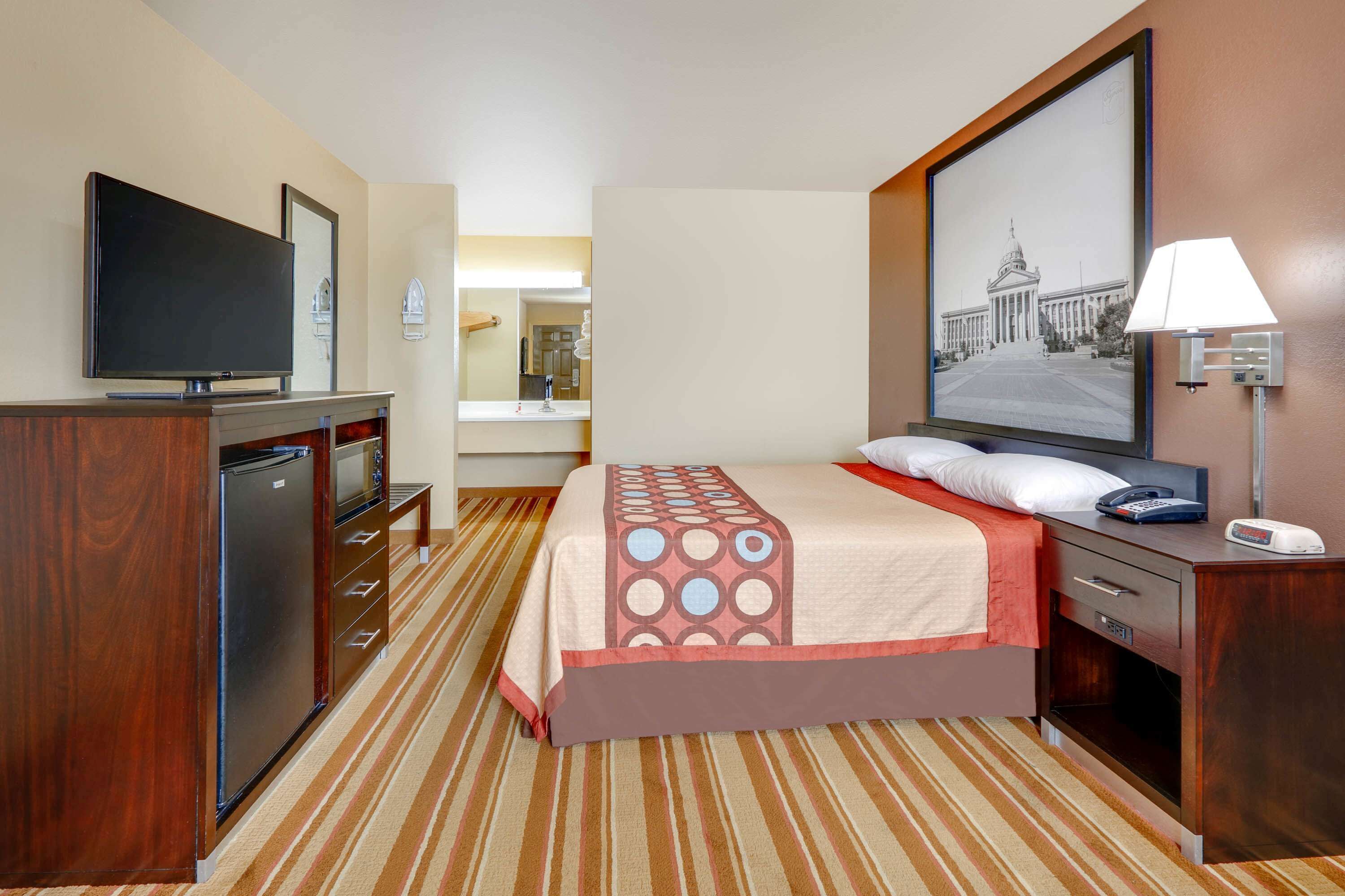 A hotel room with a bed, a TV, a desk, a lamp, and a painting on the wall.