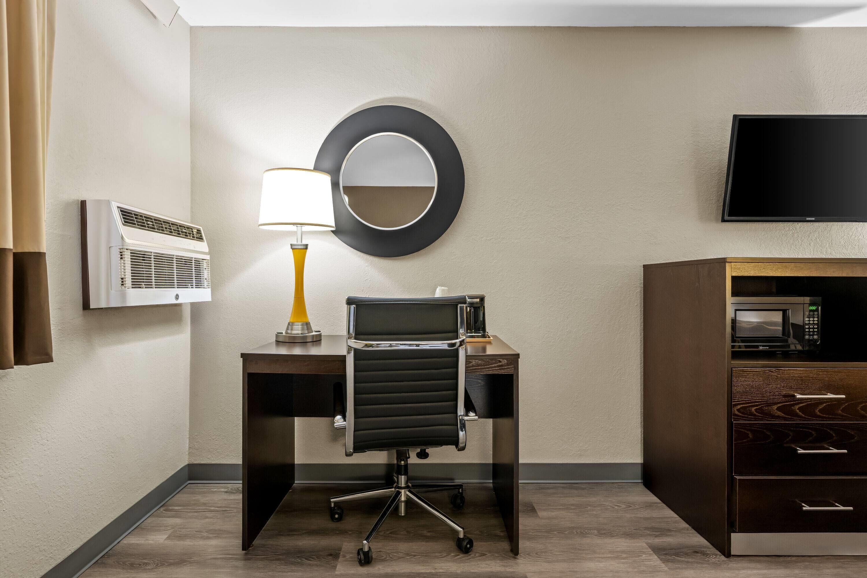 A hotel room with a desk, chair, lamp, air conditioner, TV, and a microwave.
