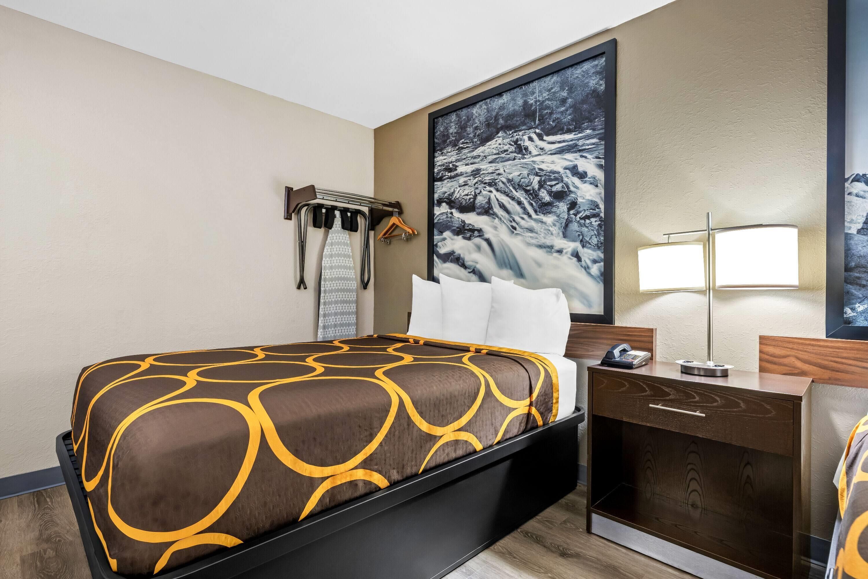 Een hotelkamer met een bed, een nachtkastje met lamp, een kapstok en een groot ingelijst schilderij aan de muur.