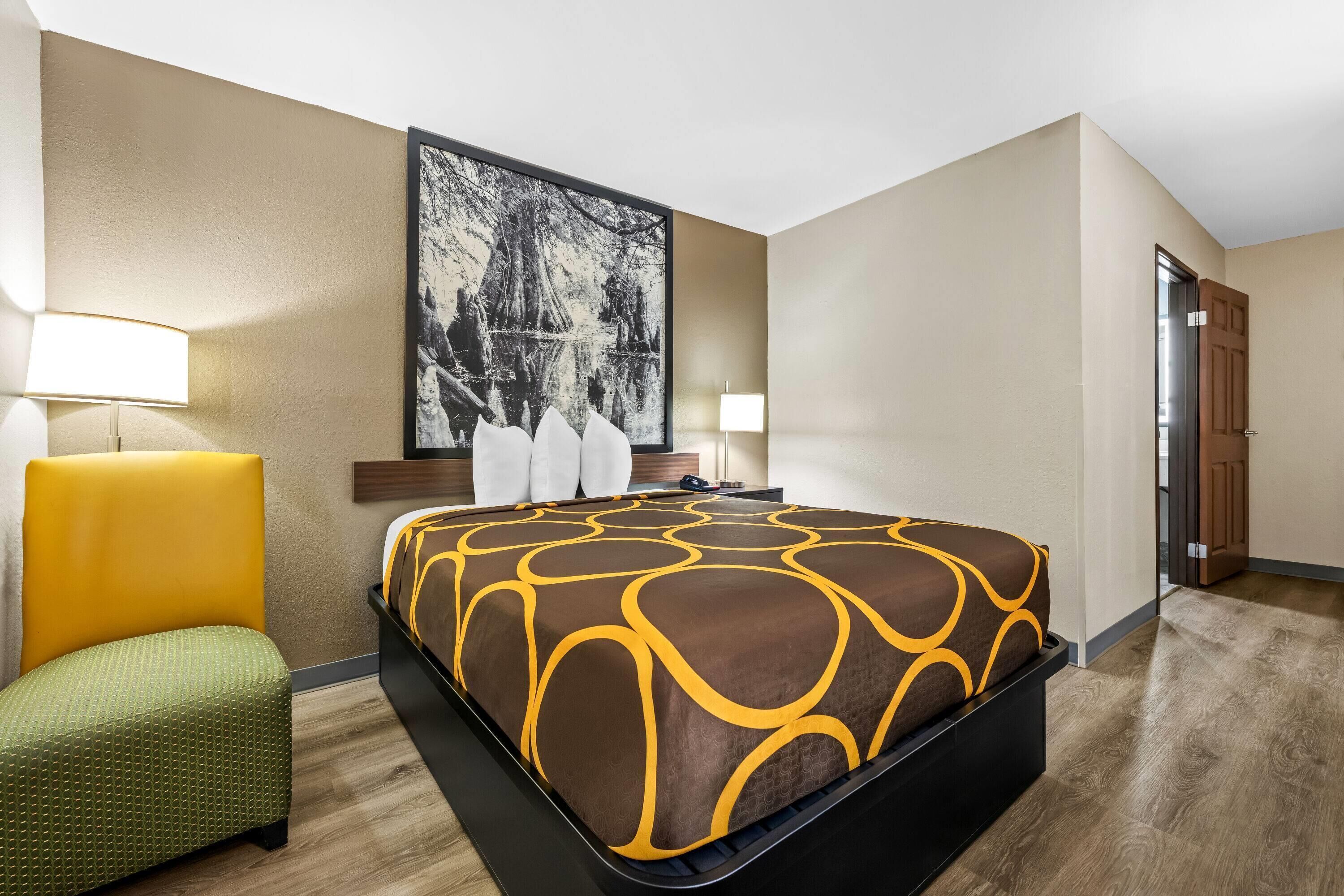A hotel room with a large bed, a green armchair, a desk with a lamp, and a large abstract painting on the wall.