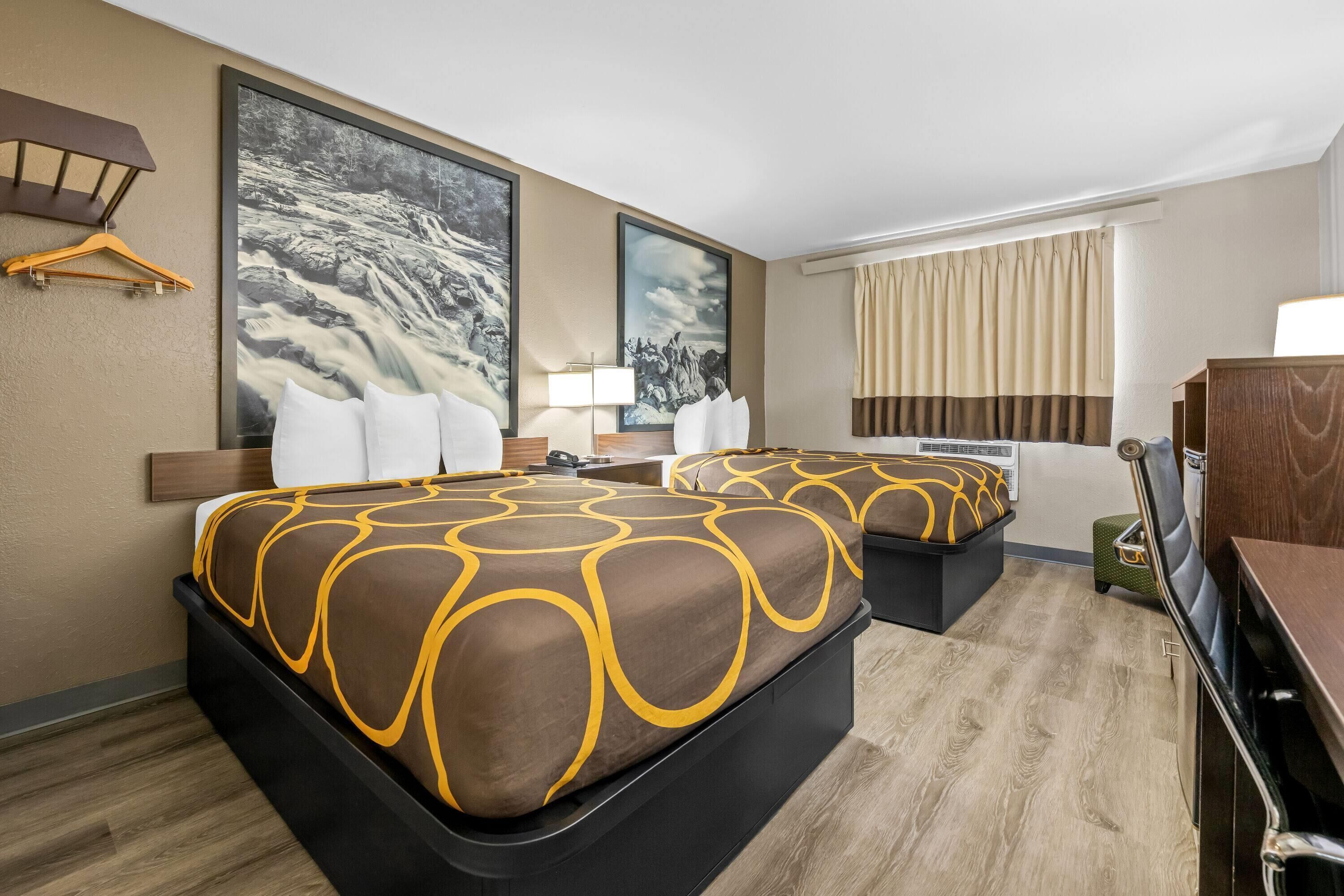 A hotel room with two beds, a wooden dresser, a desk, a hanging light fixture, and a large painting on the wall.