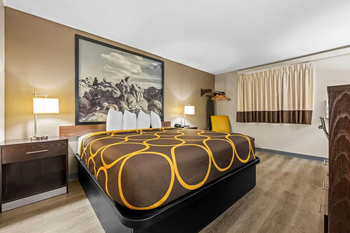 Habitación de hotel con cama, lámparas de noche, un cuadro grande en la pared, una ventana con cortinas y piso de madera.