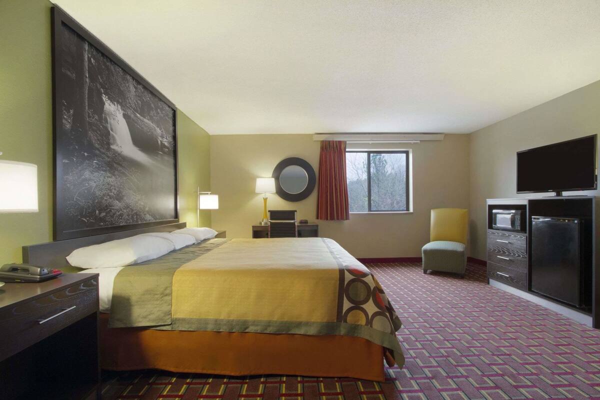 Habitación de hotel con cama, escritorio, silla, televisión, refrigerador y ventana con cortinas.