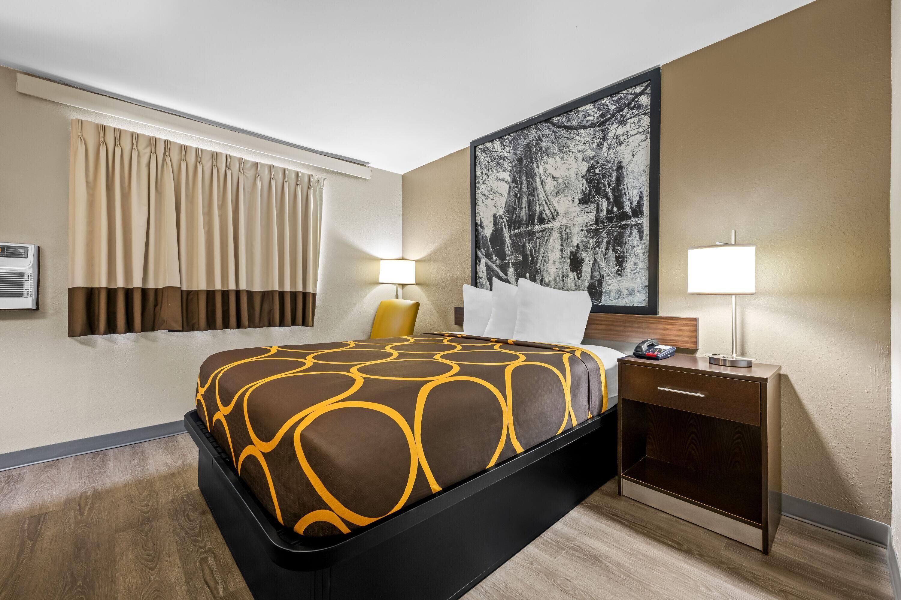 A hotel room with a bed, bedside tables, a nightstand, a wall-mounted air conditioner, and a large framed picture.