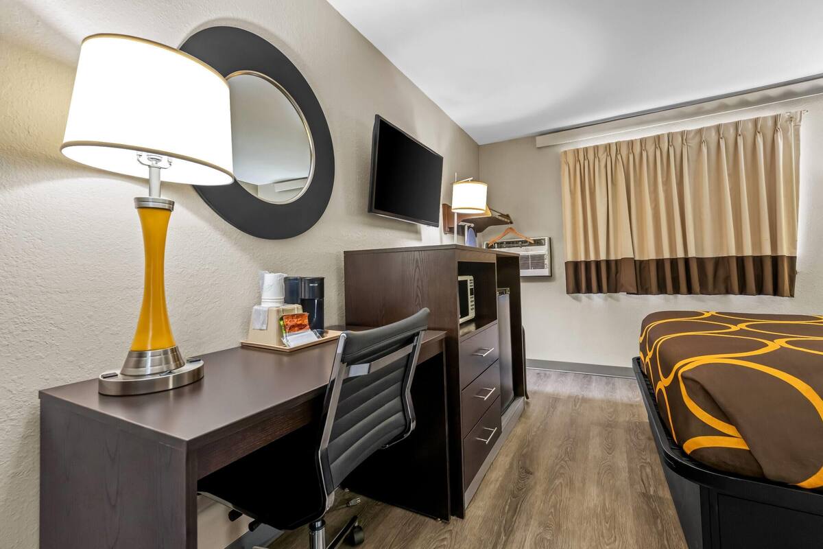 Habitación de hotel con escritorio, una silla, cama, televisión, espejo, ventana con cortinas y lámpara de pared.