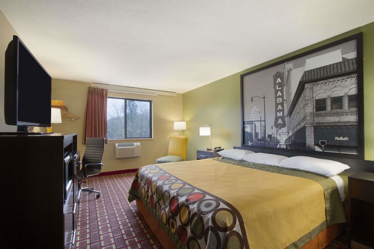 Una habitación de hotel con una cama, un escritorio con televisor, una silla, una ventana con cortinas, una lámpara y un cuadro de paisaje urbano en la pared.