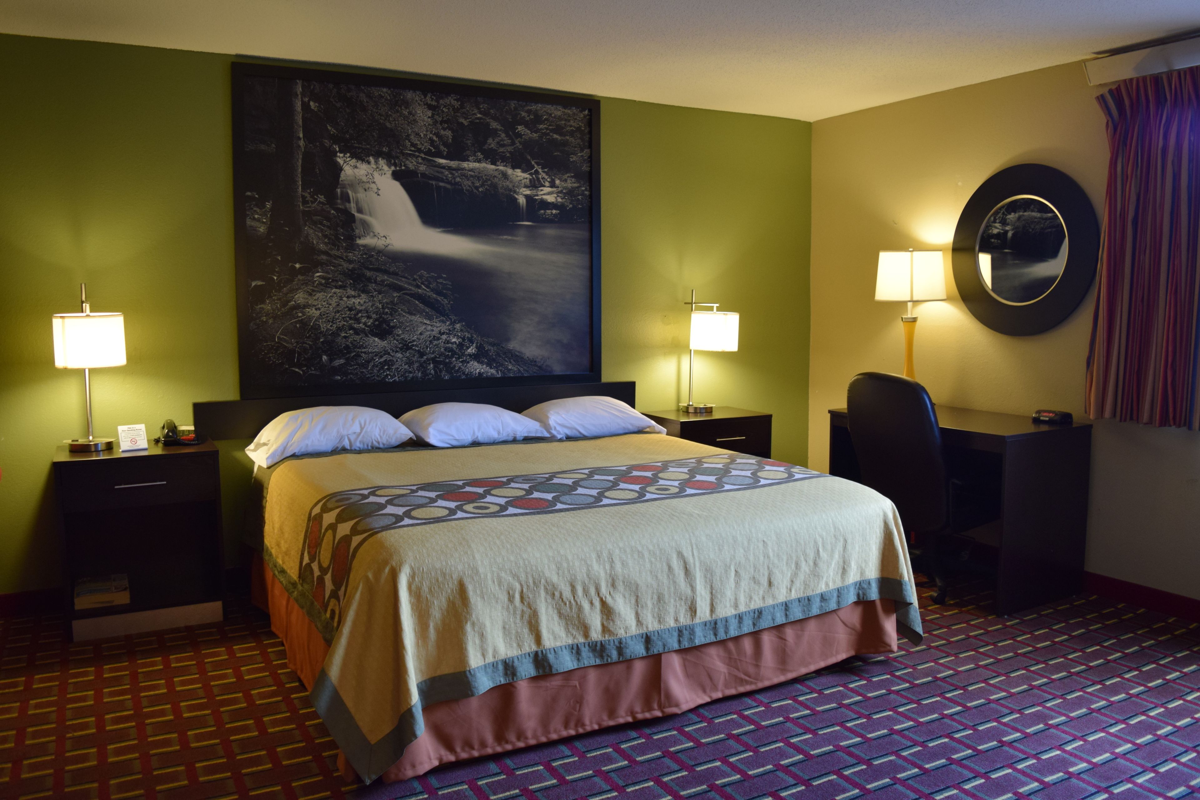 A hotel room with a large bed, two bedside tables with lamps, a desk with a chair, a large framed picture on the wall, and a patterned carpet.
