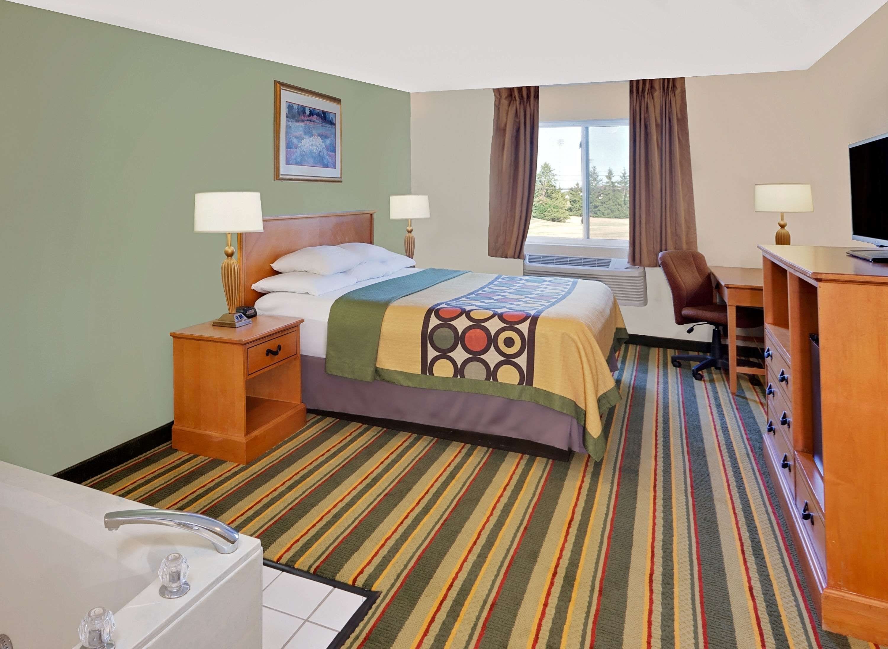 A hotel room with a bed, two bedside tables with lamps, a desk with a chair, a television, and a large window with curtains.