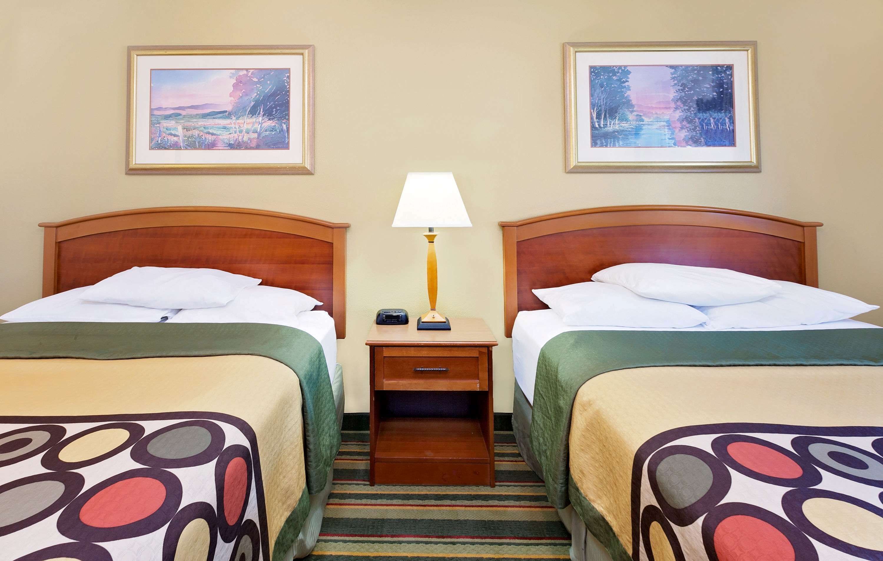 Una camera d'albergo con due letti, un comodino con una lampada e due quadri appesi al muro.