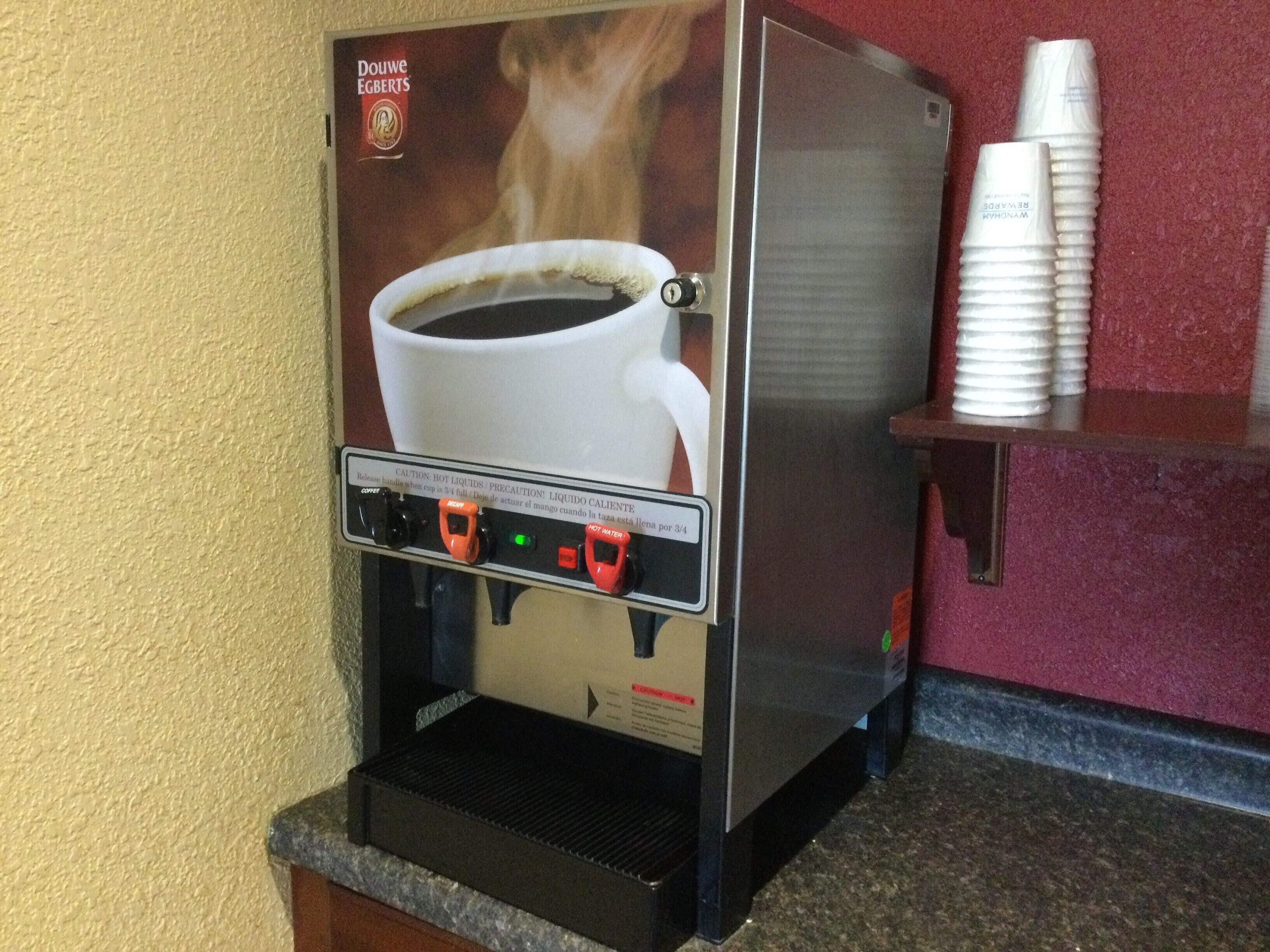 A coffee machine with a cup of coffee and steam, a control panel with buttons, and a stack of disposable cups on a counter.