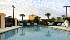 Una piscina con sillas de descanso y una valla, rodeada de palmeras y un edificio de hotel al fondo.