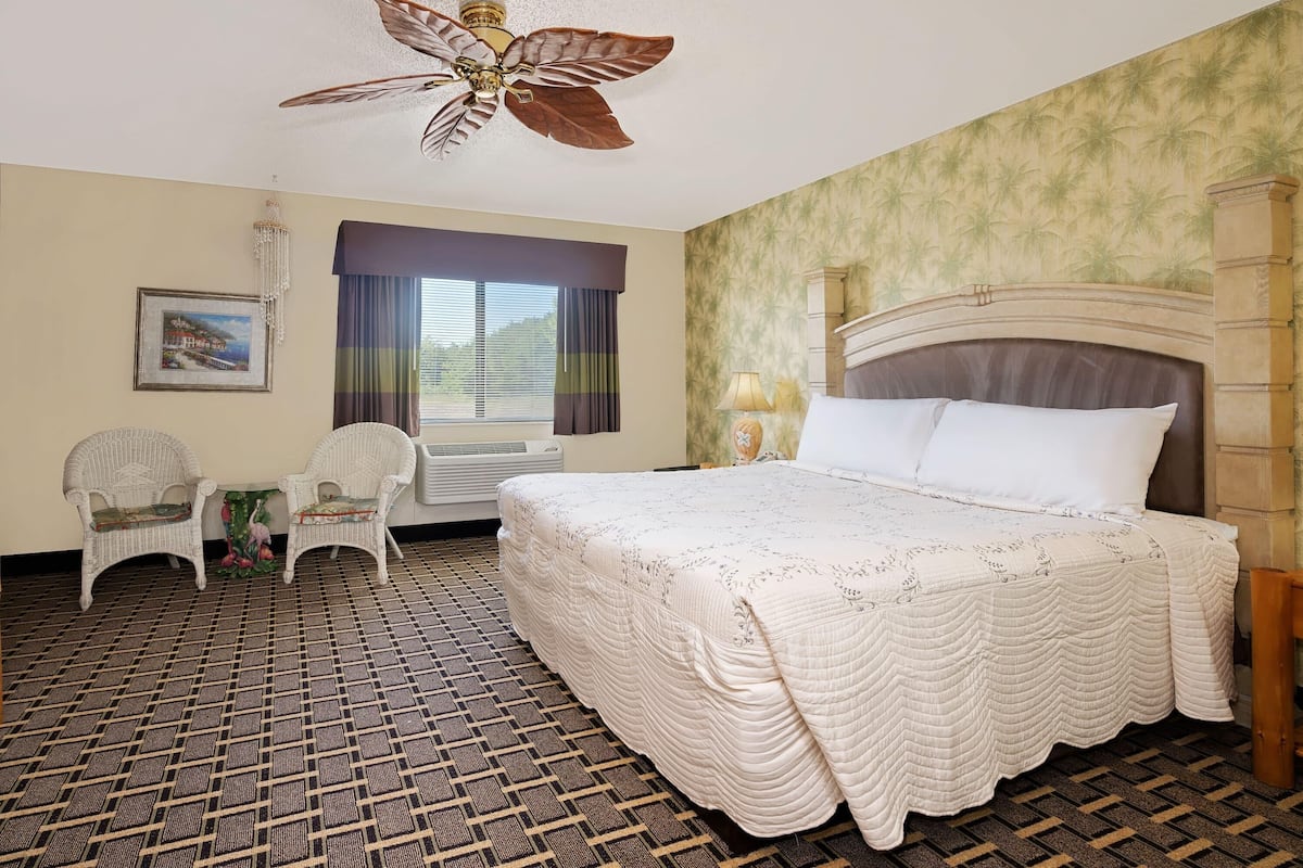 Habitación de hotel con una cama grande, dos sillas, un ventilador de techo, una ventana con cortinas y un piso estampado.