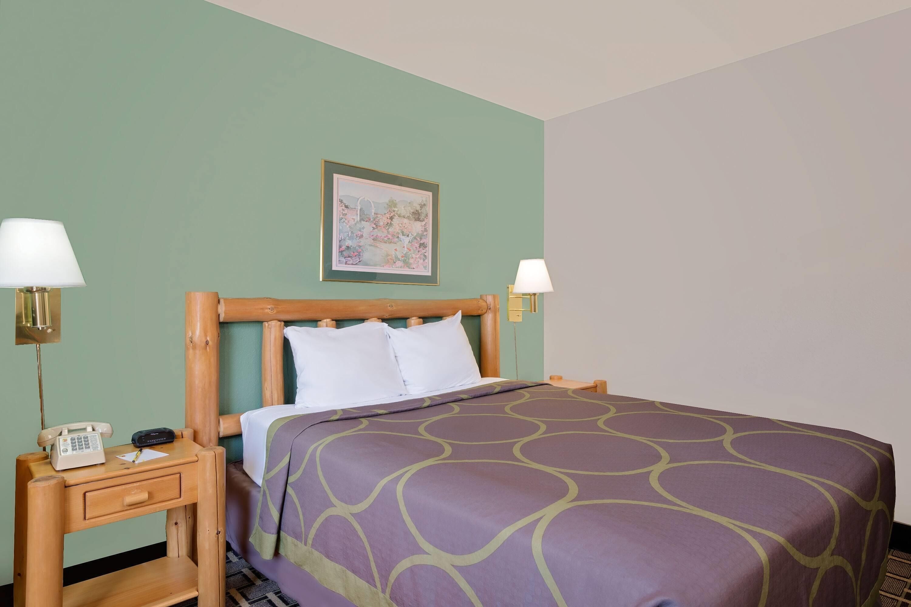 A neatly made bed with a purple and green patterned bedspread, two bedside lamps, a wooden nightstand with a telephone, and a framed picture on the wall.