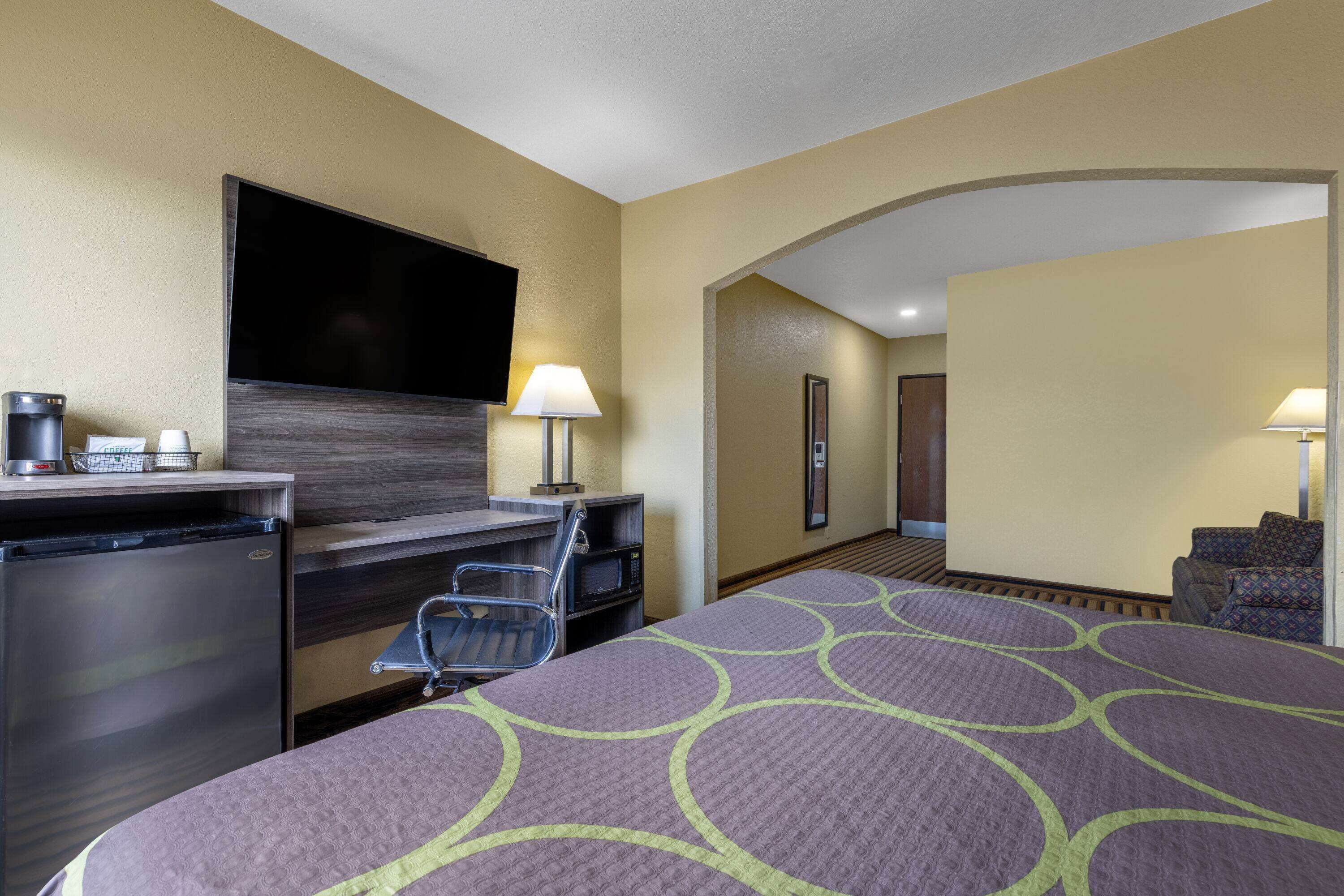 A hotel room with a bed, desk, microwave, refrigerator, TV, and a seating area with a chair and lamp.