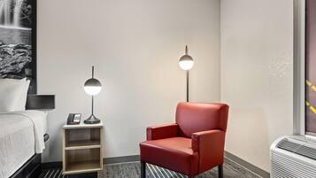 Habitación de hotel con una cama, un sillón rojo, una mesita de noche con lámpara y un calentador de pared.