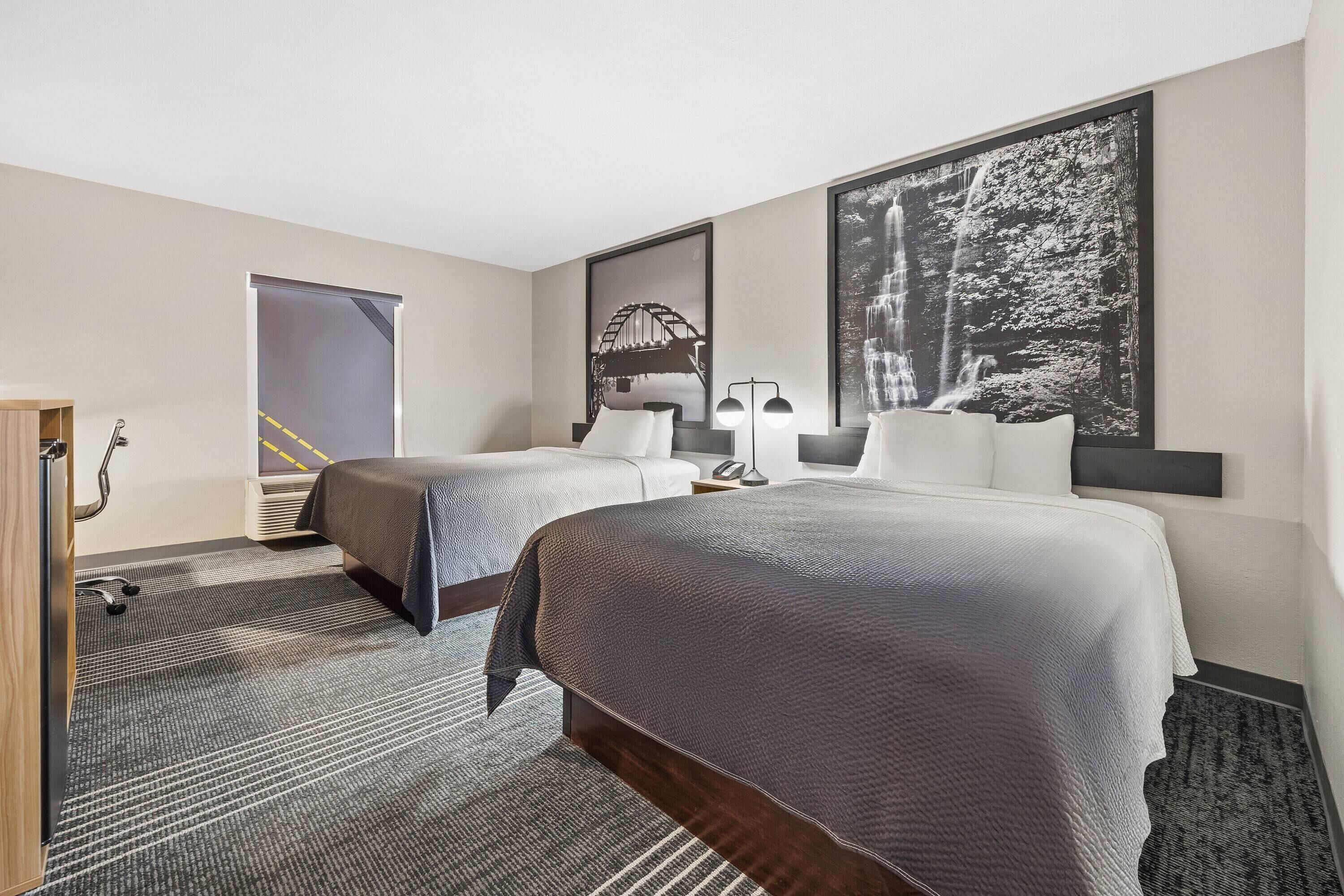 Une chambre d’hôtel avec deux lits, un bureau, une chaise, une lampe et un grand tableau encadré au mur.