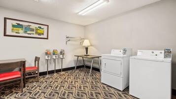 Una habitación de hotel pequeña con alfombra estampada, un banco de madera con cojines rojos, una mesa, una lámpara, un soporte con artículos de aseo y dos lavadoras blancas.