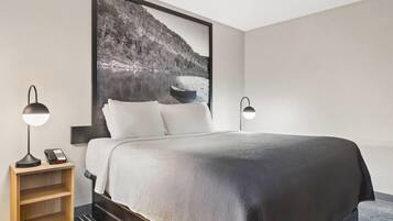 A neatly made bed with a large framed picture of a lake and mountains, two bedside lamps, a wooden nightstand with a phone, and a small shelf.