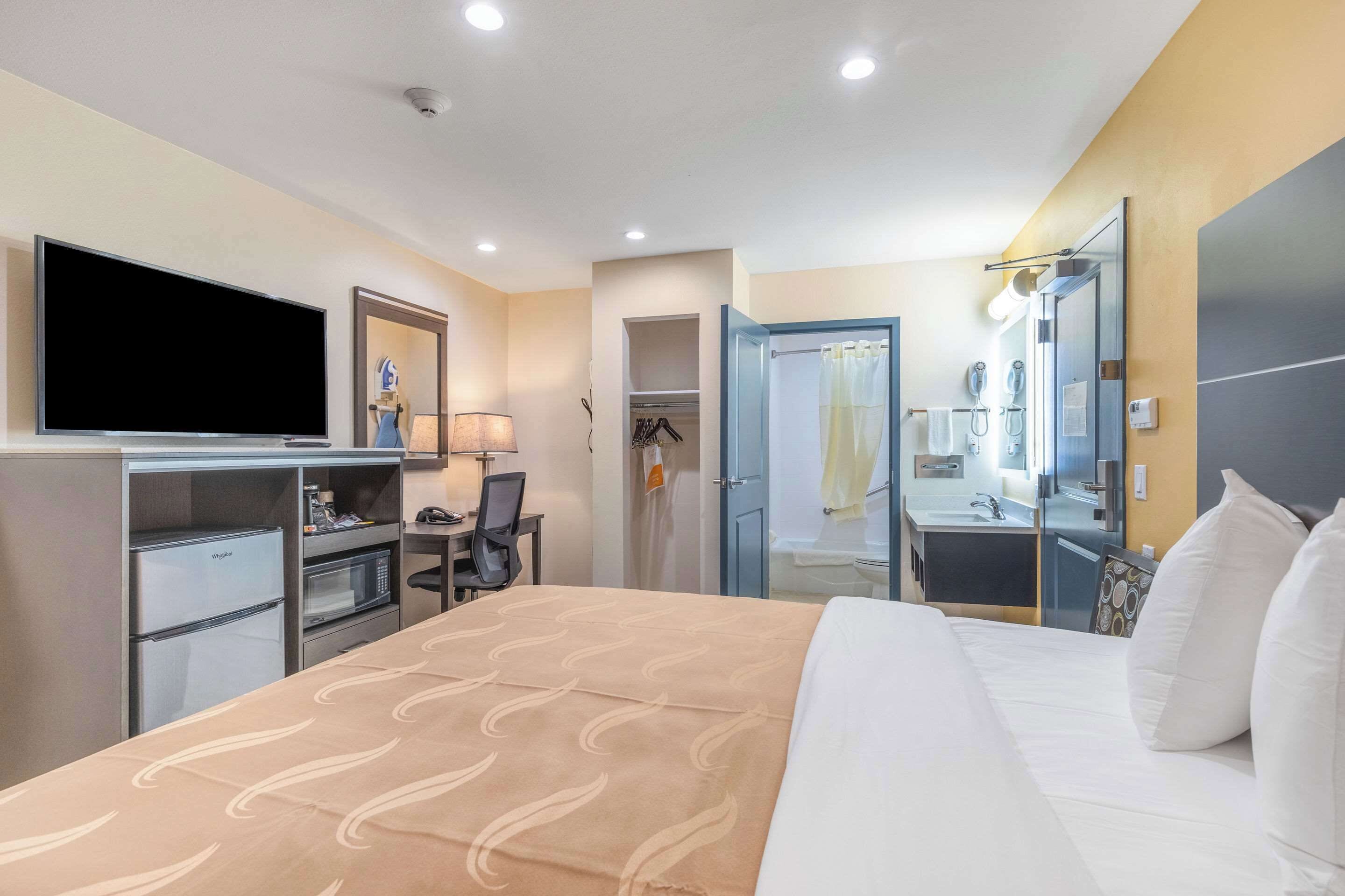 A hotel room with a bed, a desk with a flat-screen TV, a microwave, a refrigerator, a shower, and a bathroom with a mirror and sink.