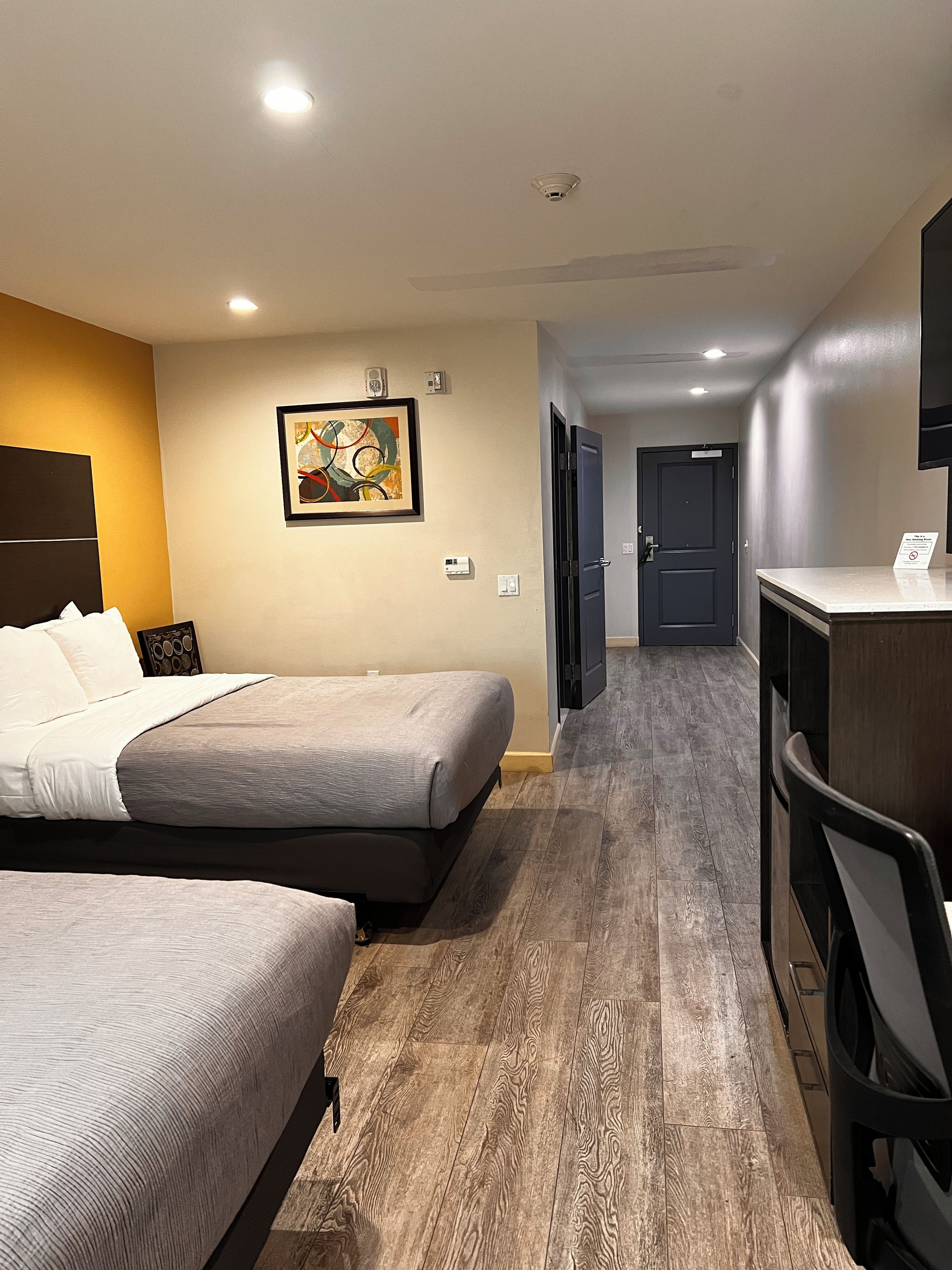 Ein Hotelzimmer mit zwei Betten, einem Schreibtisch, einem Stuhl, einem Bild an der Wand und einer Tür zu einem anderen Raum.